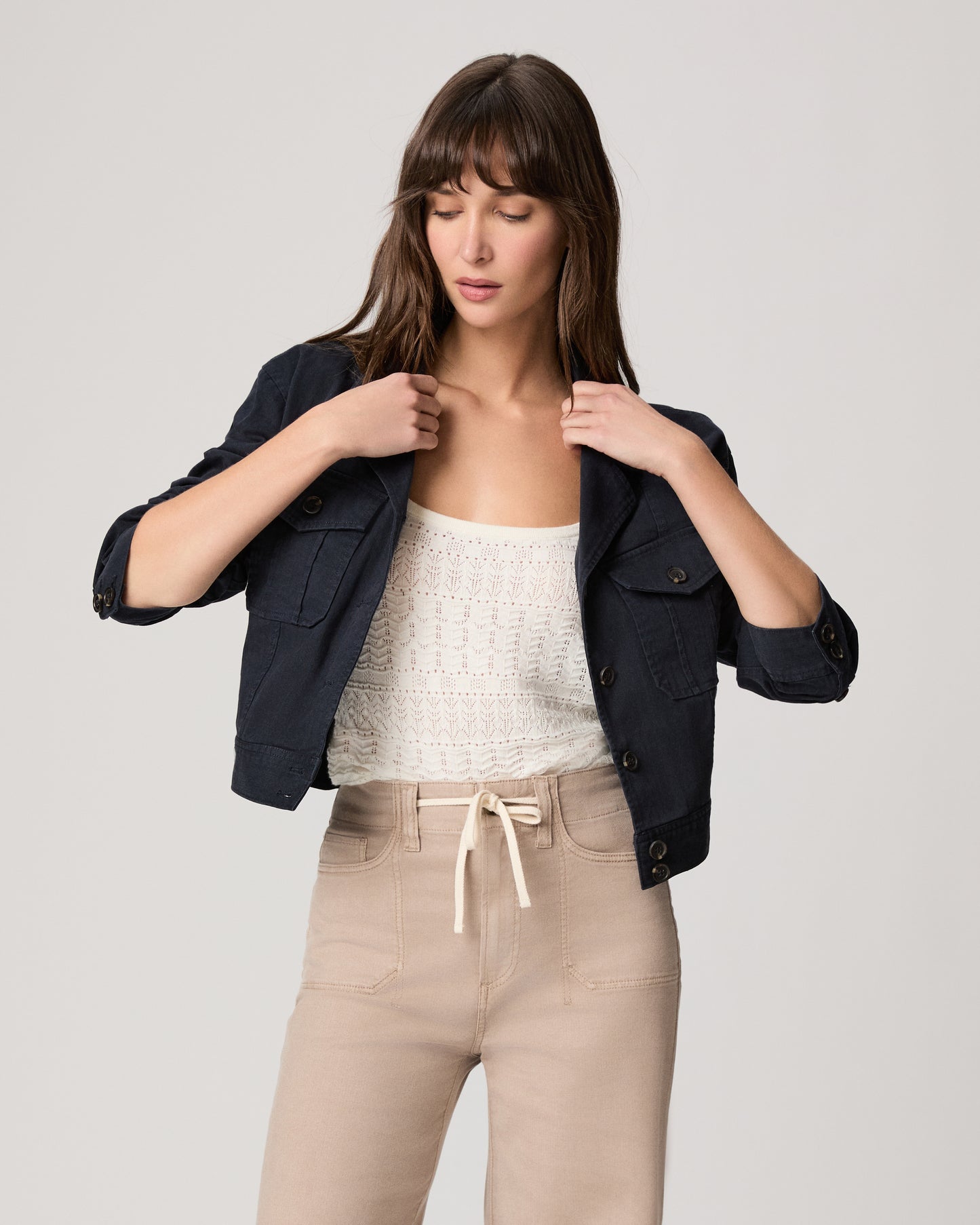 Woman wearing a navy jacket over a white top and beige pants on a light gray background
