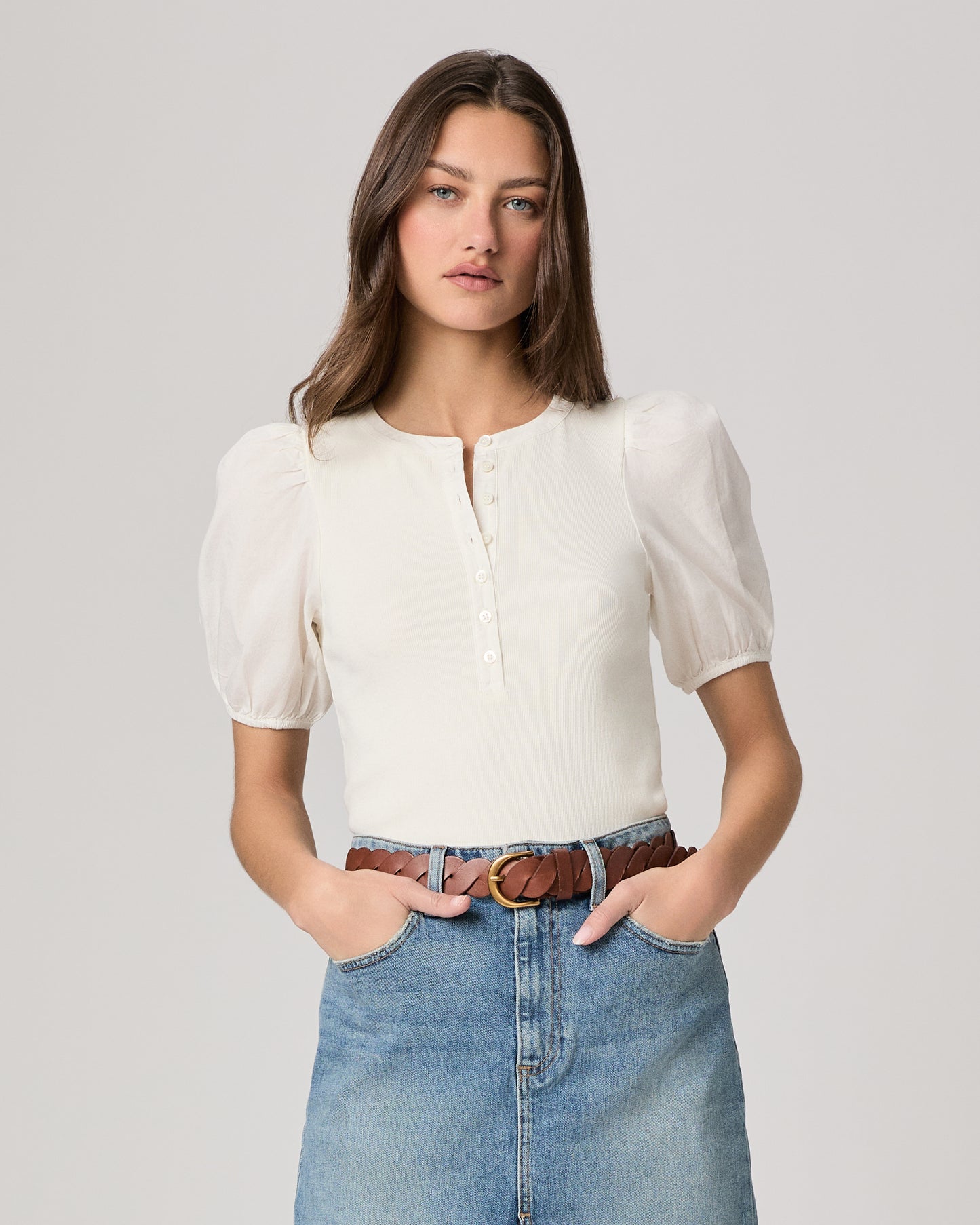 Woman wearing a white blouse and blue denim skirt paired with brown belt on a plain background