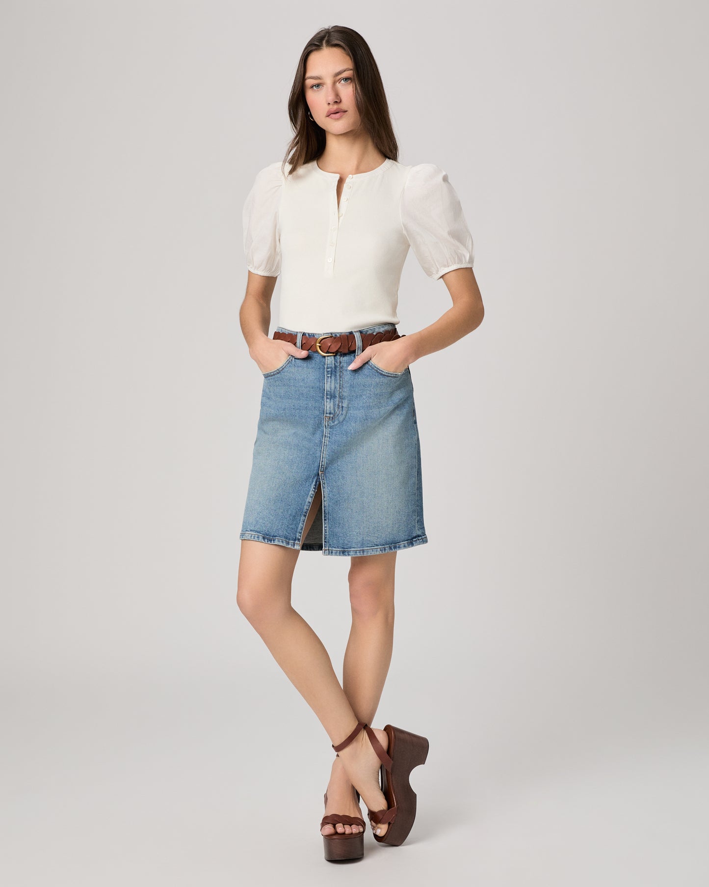 Woman wearing a white blouse and blue denim skirt paired with brown belt and heels on a plain background
