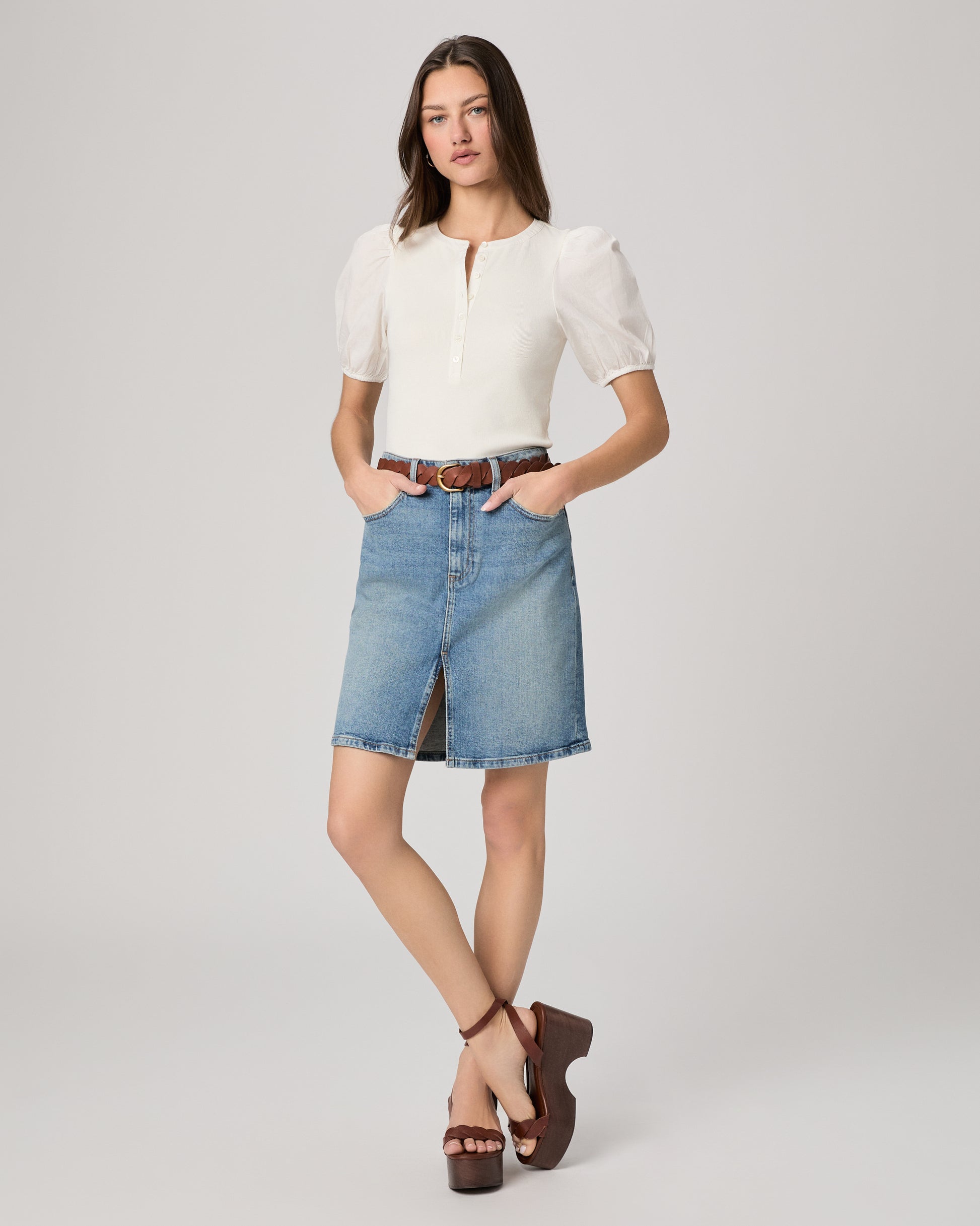 Woman wearing a white blouse and blue denim skirt paired with brown belt and heels on a plain background