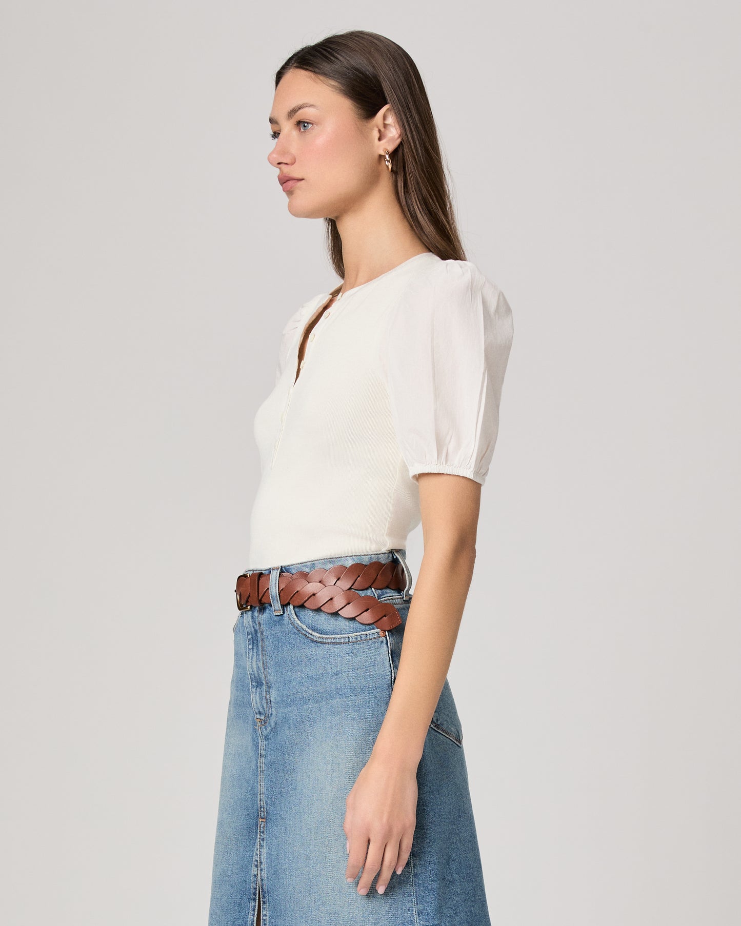 Woman wearing a white blouse and blue denim skirt paired with brown belt on a plain background