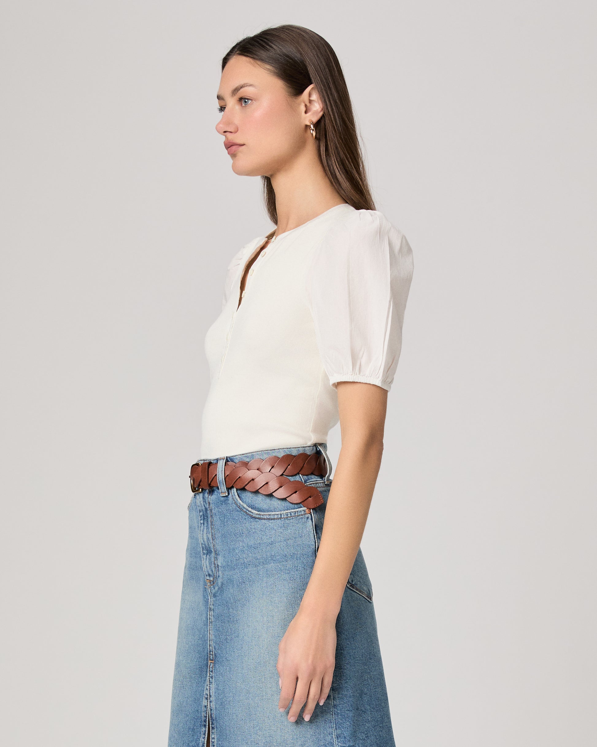 Woman wearing a white blouse and blue denim skirt paired with brown belt on a plain background