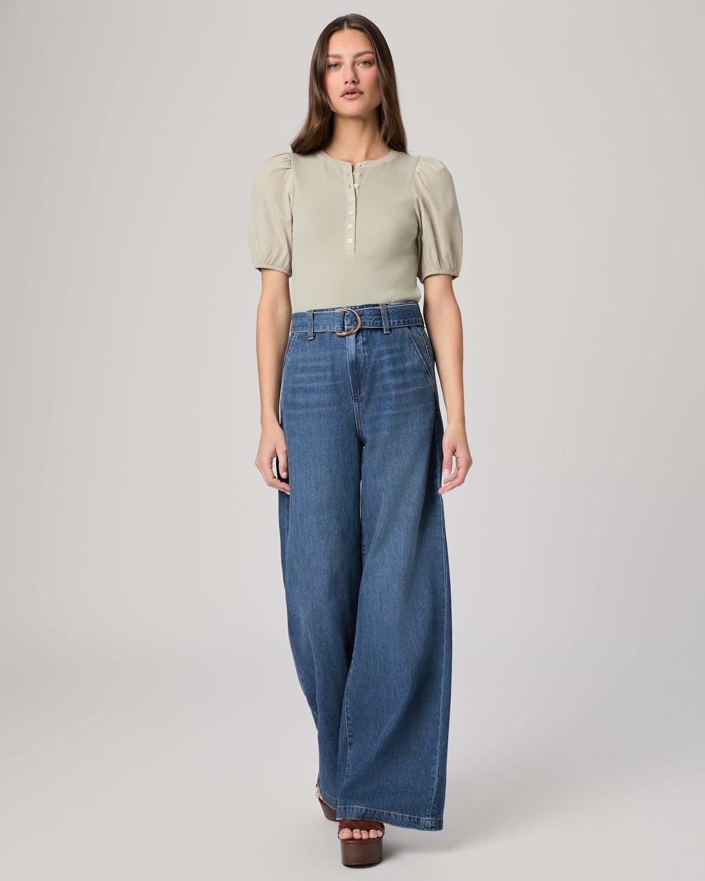 Woman wearing a light green blouse with puffed sleeves and blue jeans paired with brown heels on a plain background