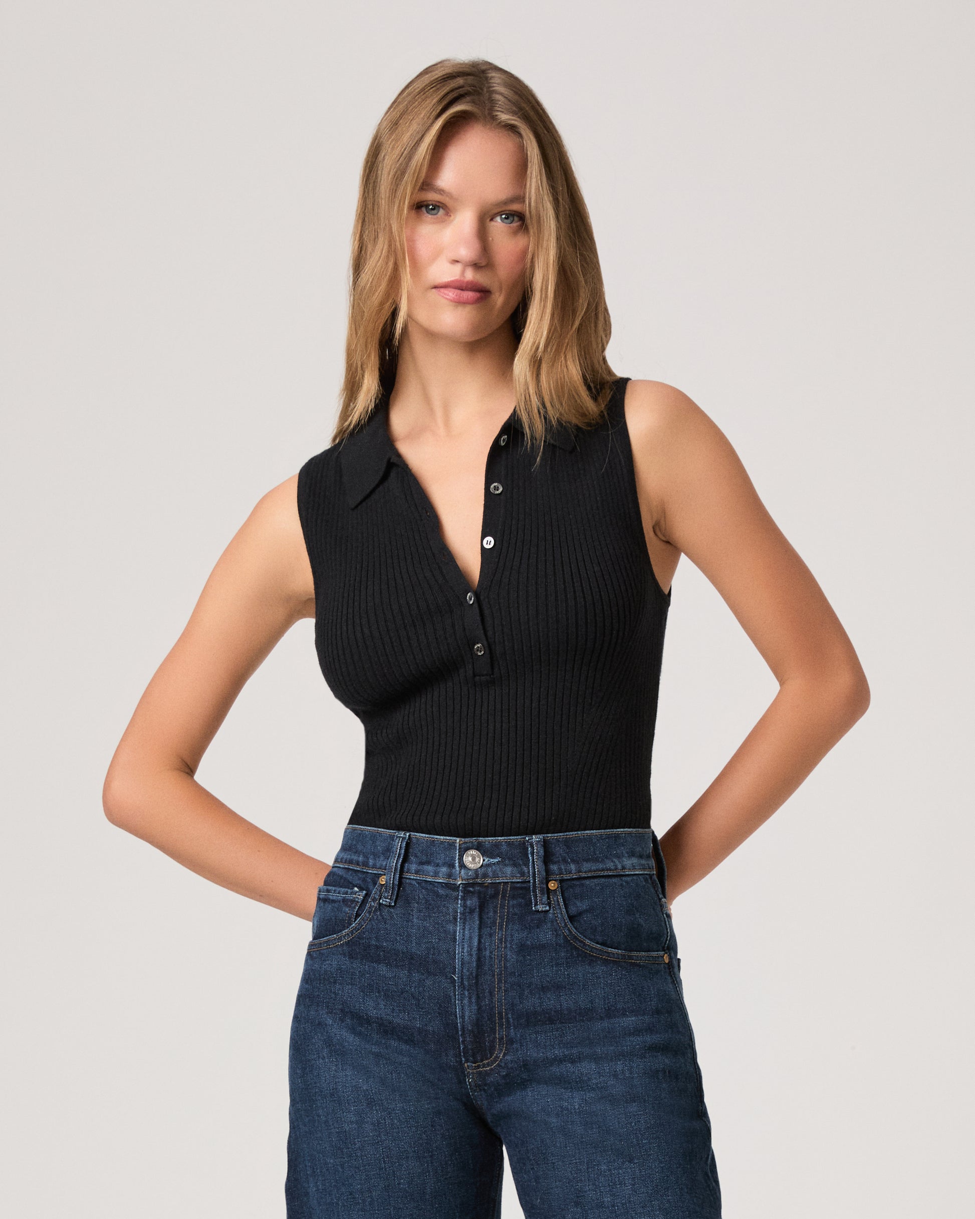 Woman wearing a black sleeveless ribbed top and blue jeans on a plain background