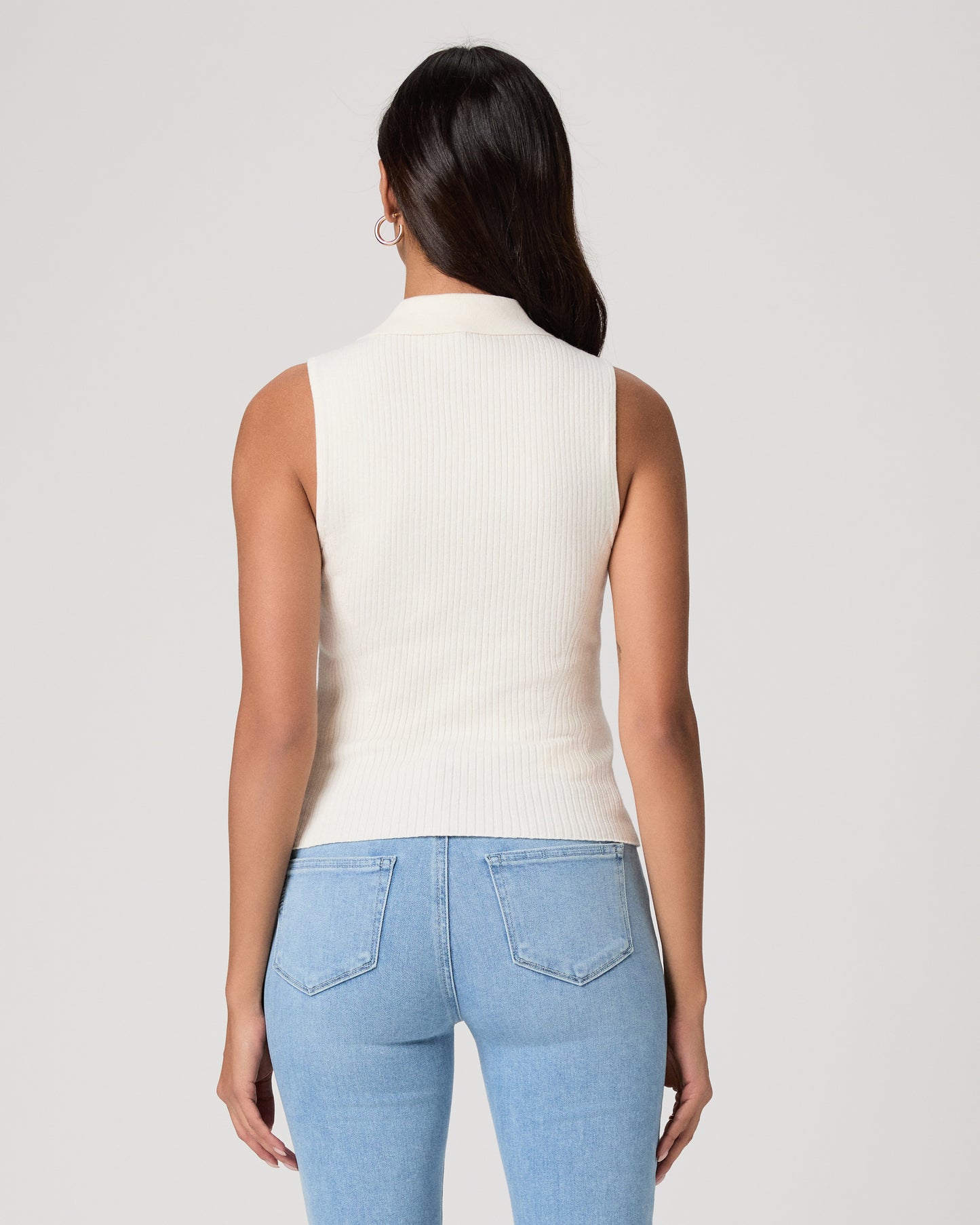 Woman wearing a white sleeveless shirt and light jeans against a white background