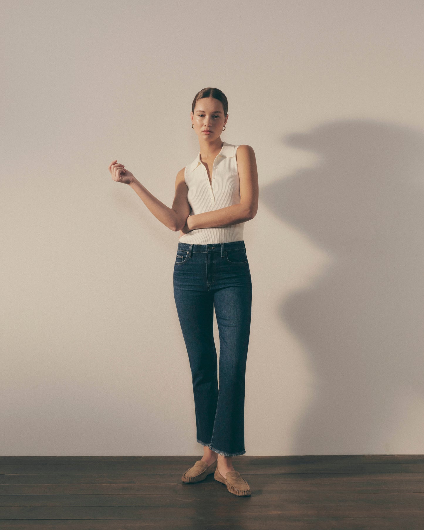 Woman wearing a white sleeveless top and dark blue jeans against a plain background