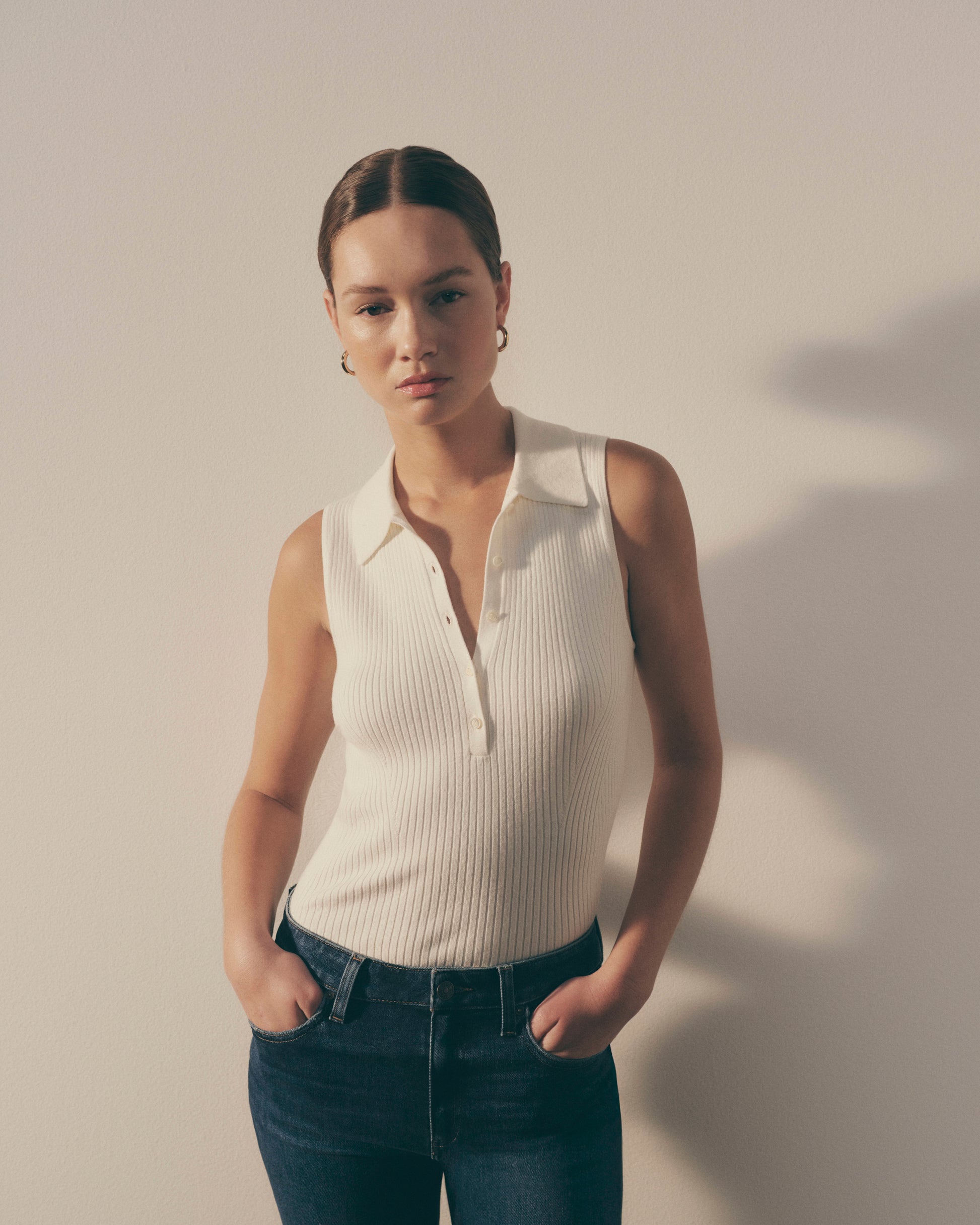 Woman wearing a white sleeveless shirt and dark jeans against a plain background