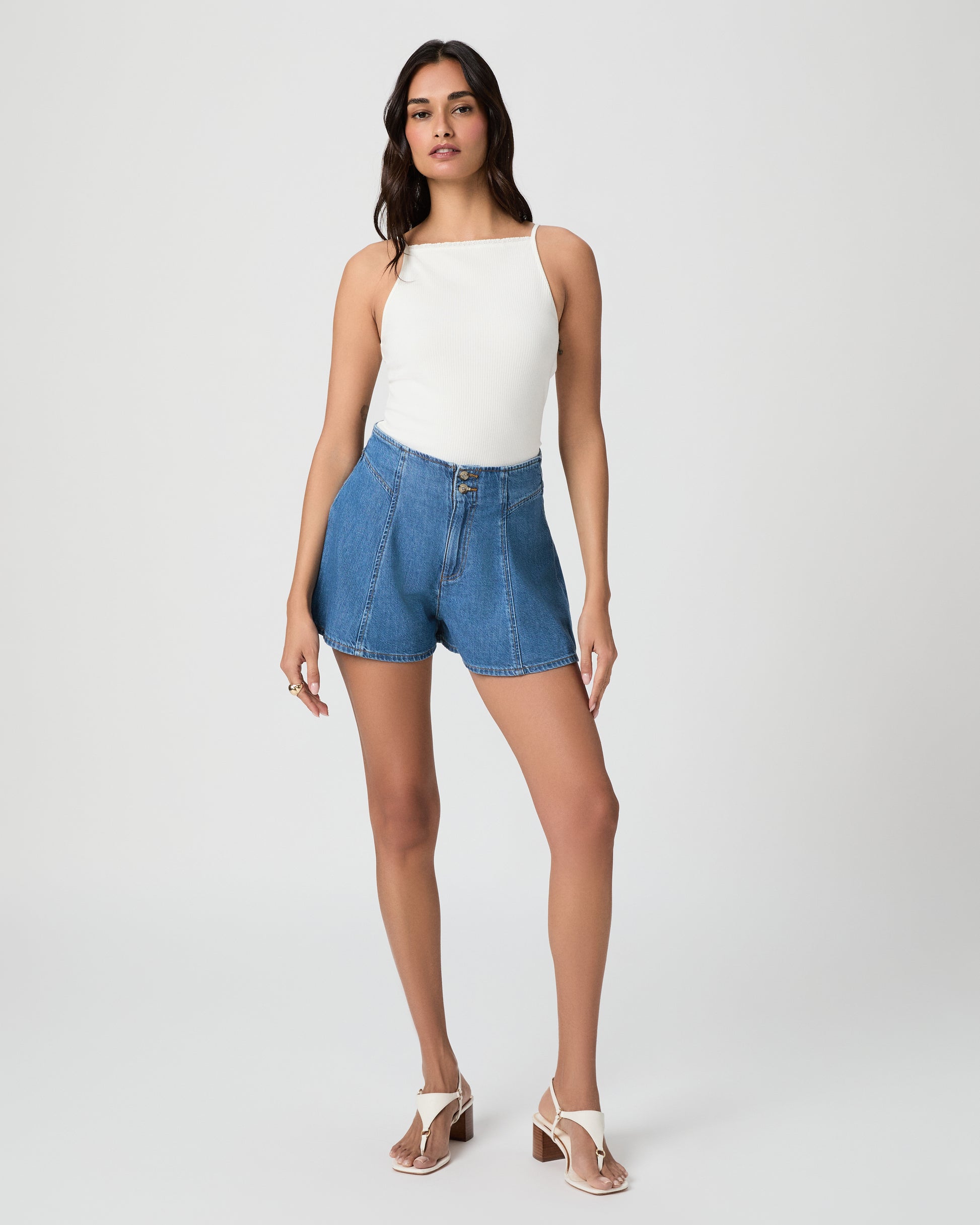 Woman wearing a white tank top and blue denim shorts paired with white sandals on a plain background