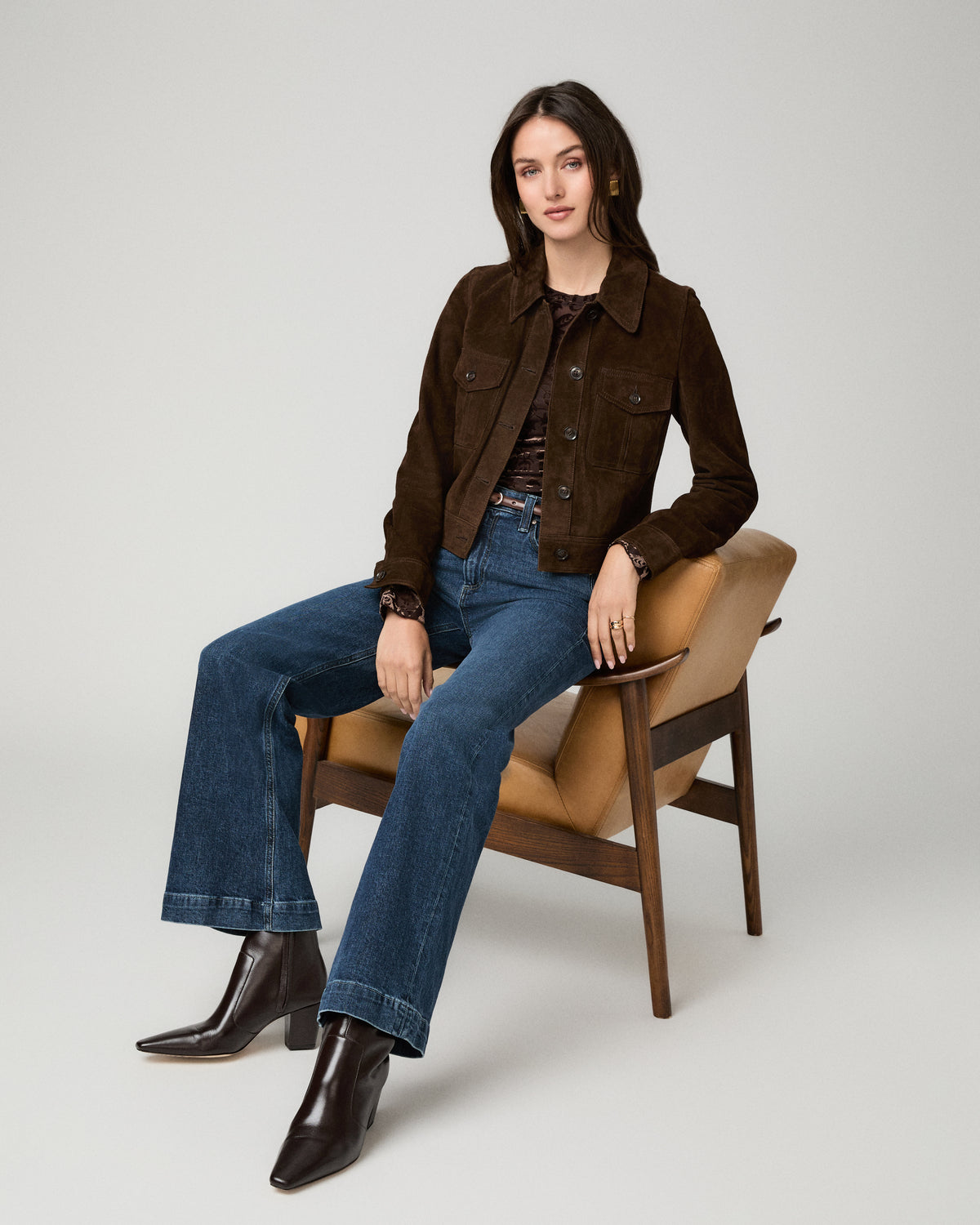 A woman sits on a mid-century modern chair wearing a dark brown suede jacket, blue flare jeans, and dark brown leather ankle boots against a light gray studio background.
