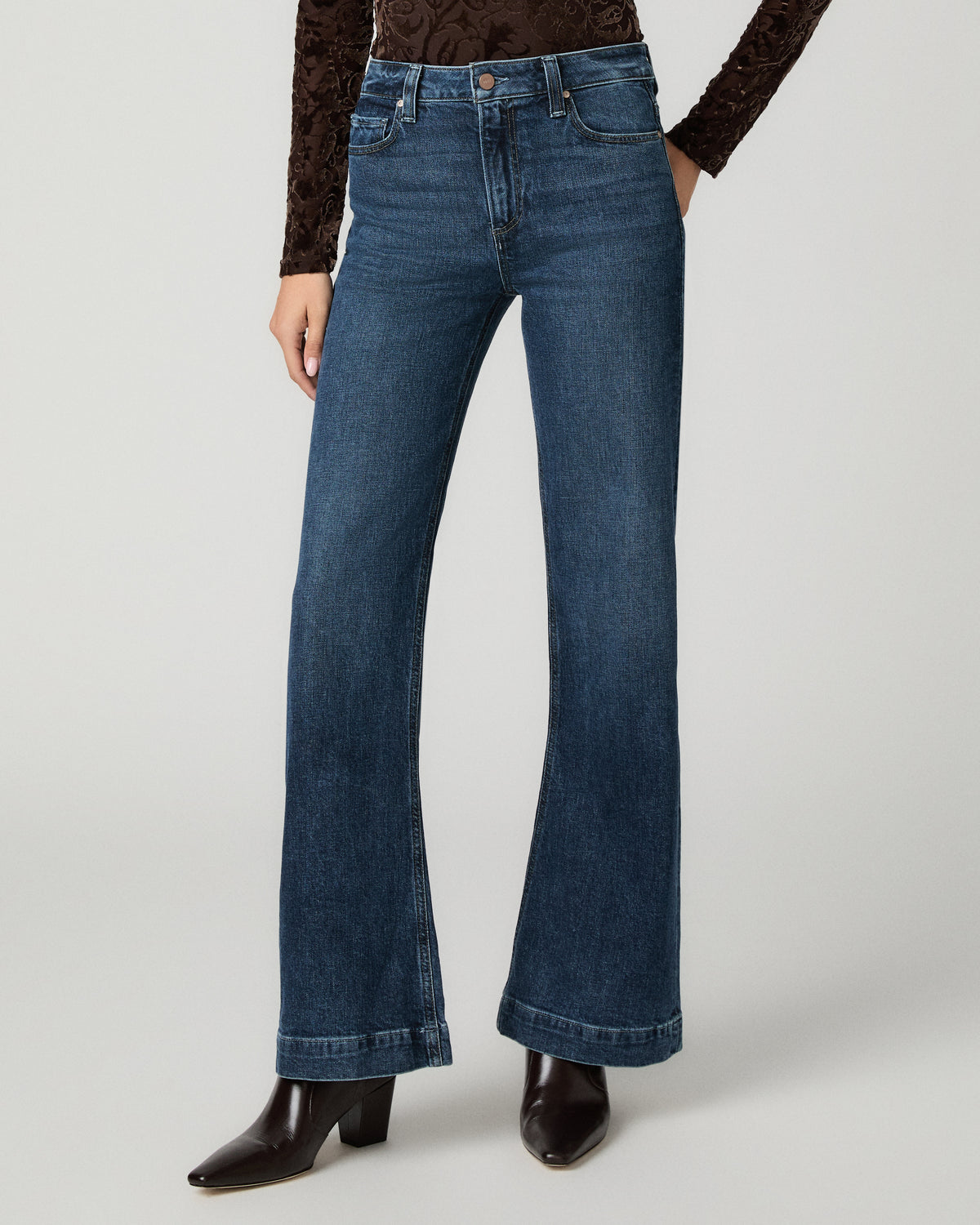A model wearing a pair of dark blue wide-leg flare jeans with a brown lace long-sleeve top and dark brown pointed-toe heeled boots, photographed against a light gray studio background.