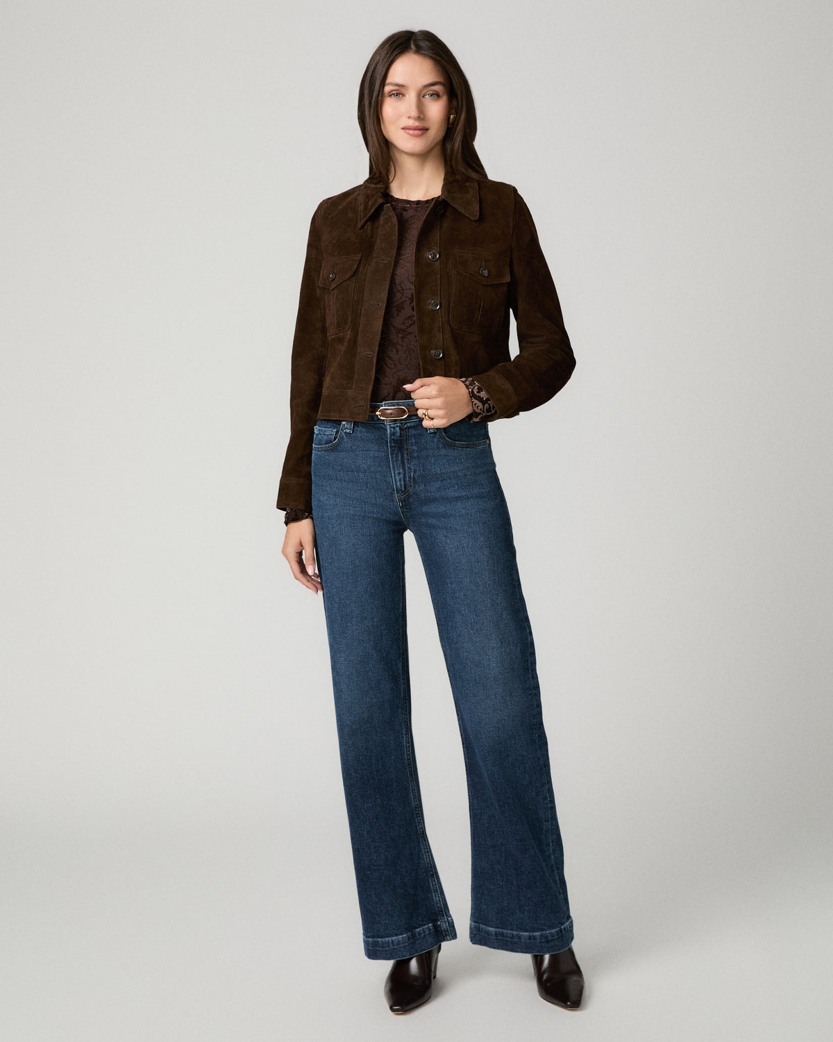 A woman stands against a light gray studio background wearing a dark brown suede jacket over a dark top, blue wide-leg jeans, and dark brown boots.