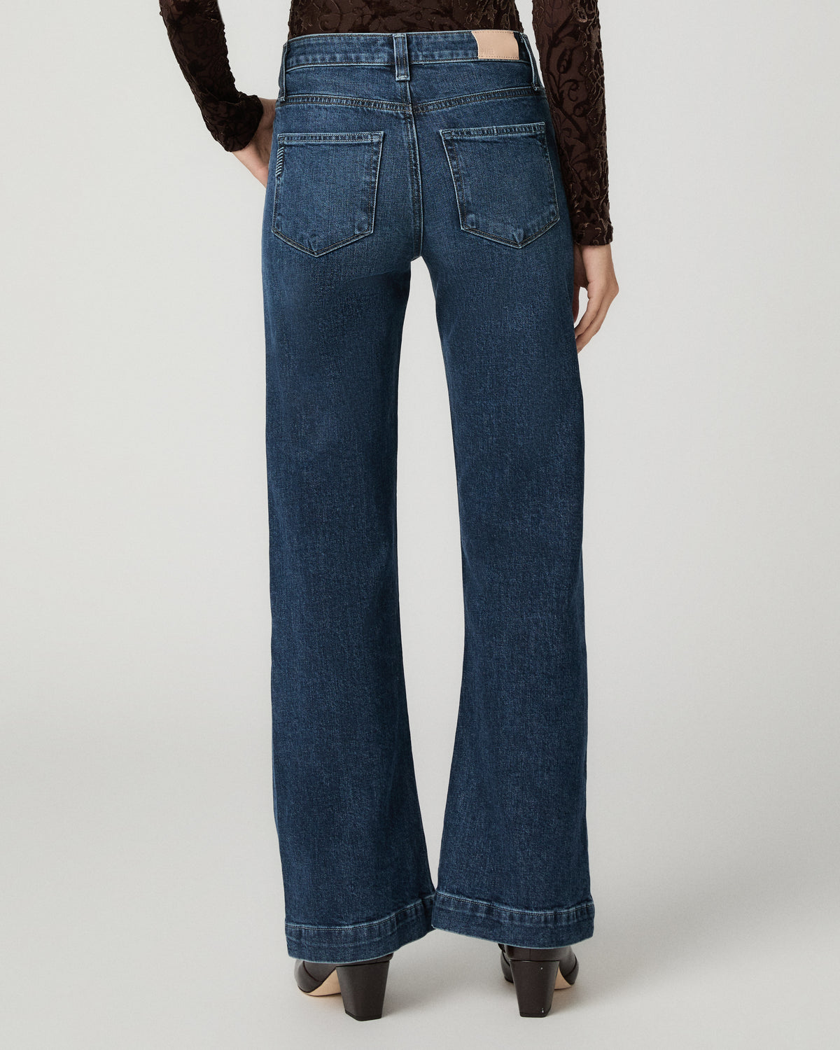 A person shown from the back wearing dark blue wide-leg denim jeans, a dark brown lace long-sleeve top, and black heels against a light gray studio background.