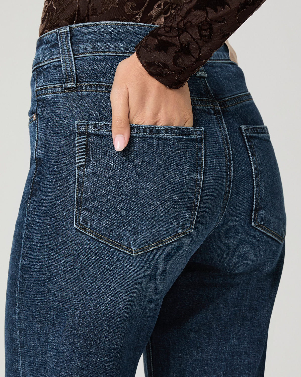 Close-up back view of a person wearing dark blue denim jeans, with one hand tucked into the back pocket. A dark brown lace long-sleeve top is partially visible at the top against a light gray studio background.
