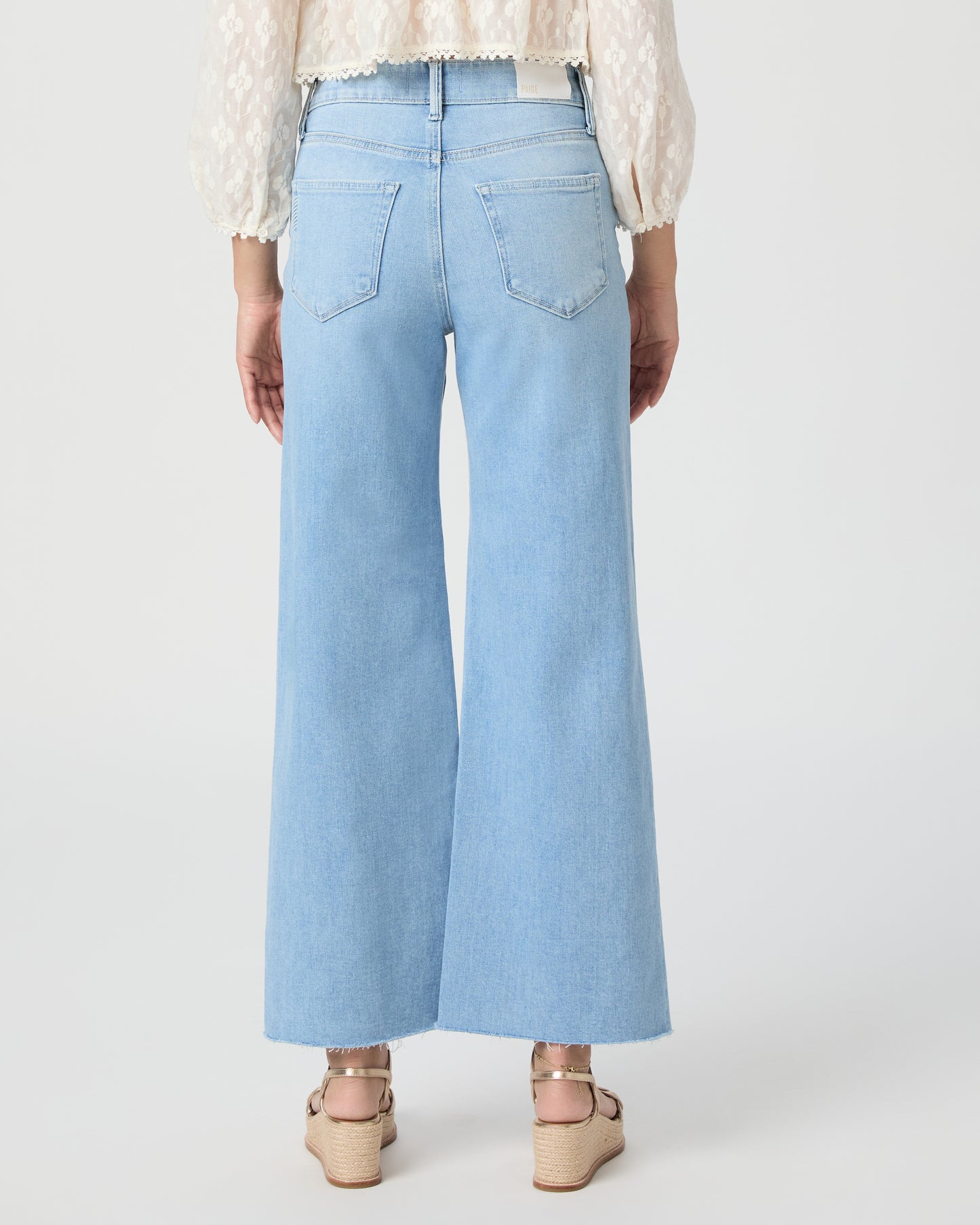 Woman wearing a white blouse and light blue jeans paired with gold wedges on a white background