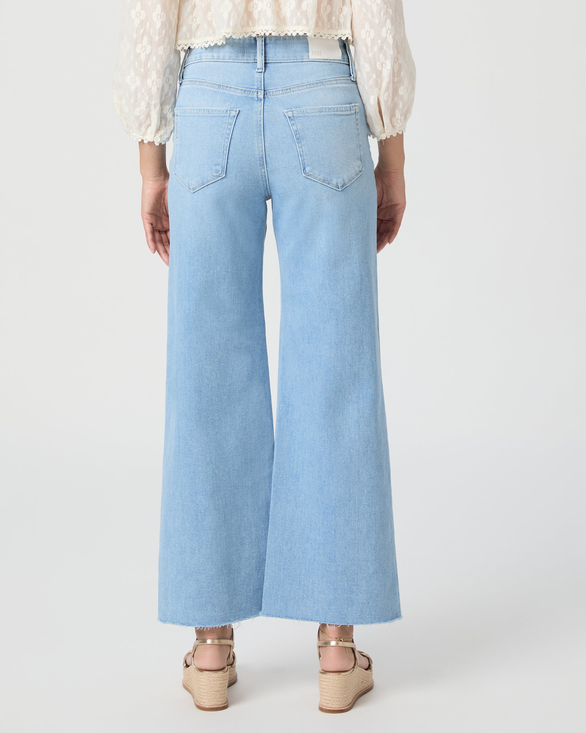 Woman wearing a white blouse and light blue jeans paired with gold wedges on a white background