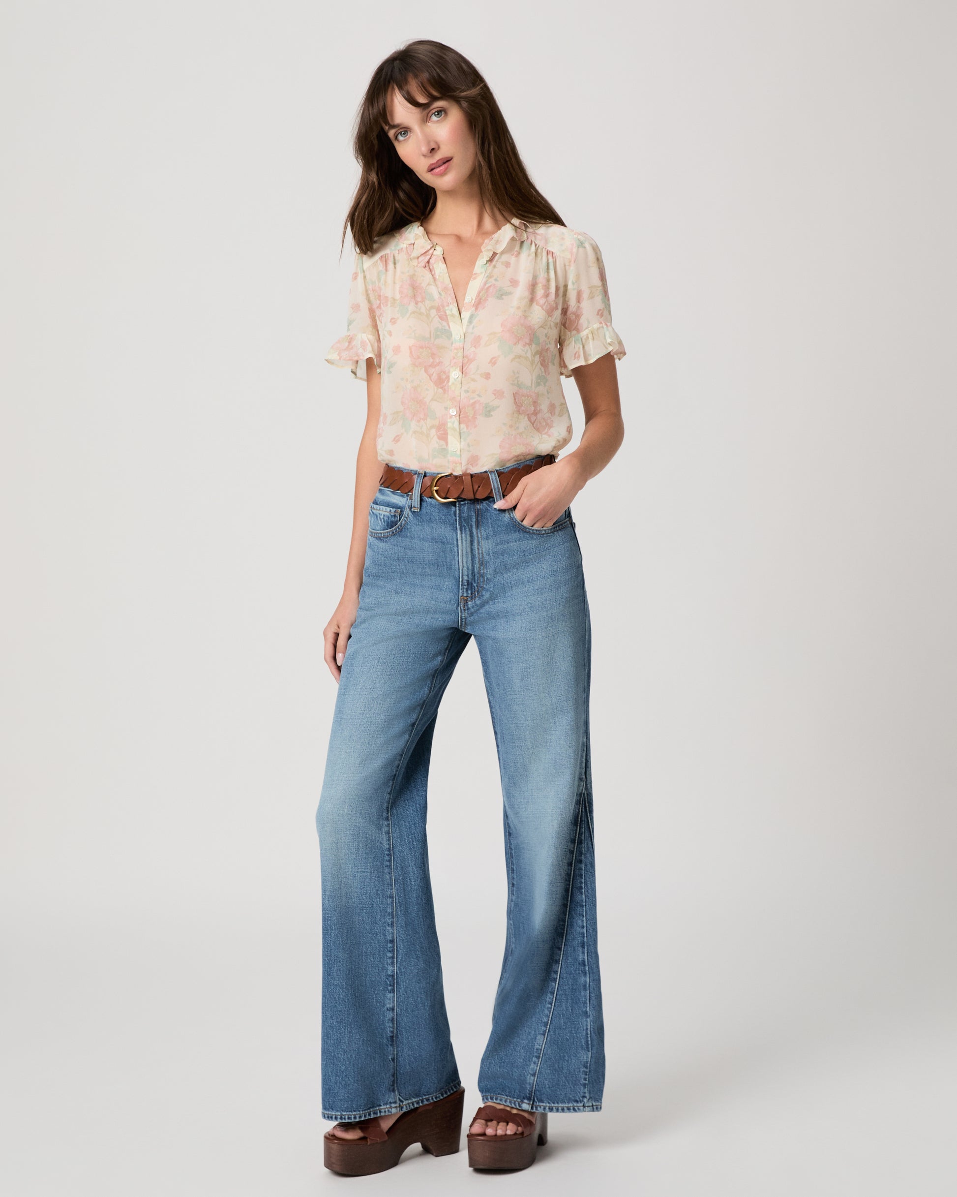 Woman wearing a flower printed light pink blouse and blue jeans paired with brown wedges  and a brown belt on a white plain background