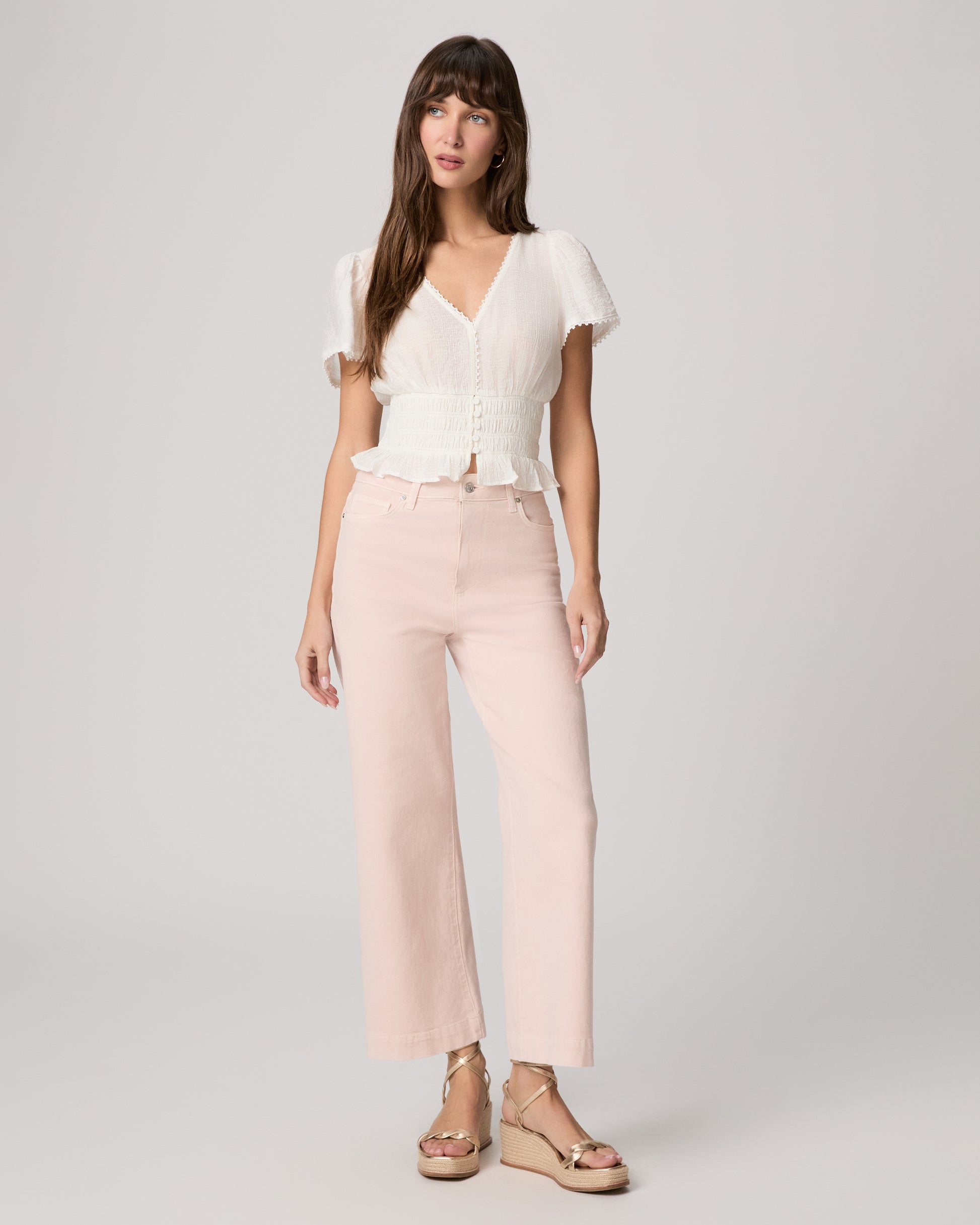 Woman wearing a white blouse and light pink pants paired with gold wedges against a plain background