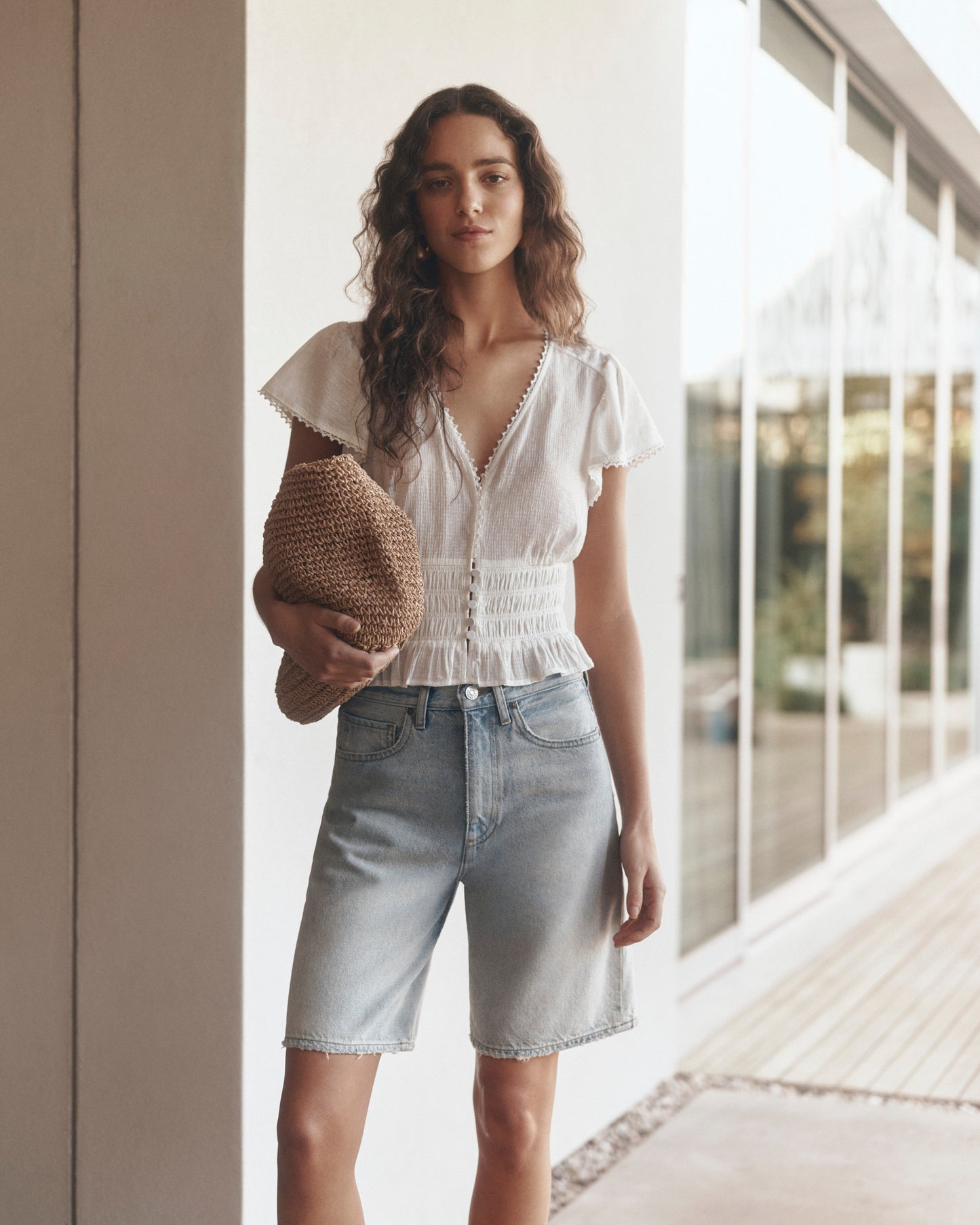 Woman wearing a white blouse and denim shorts holding a brown bag indoors.