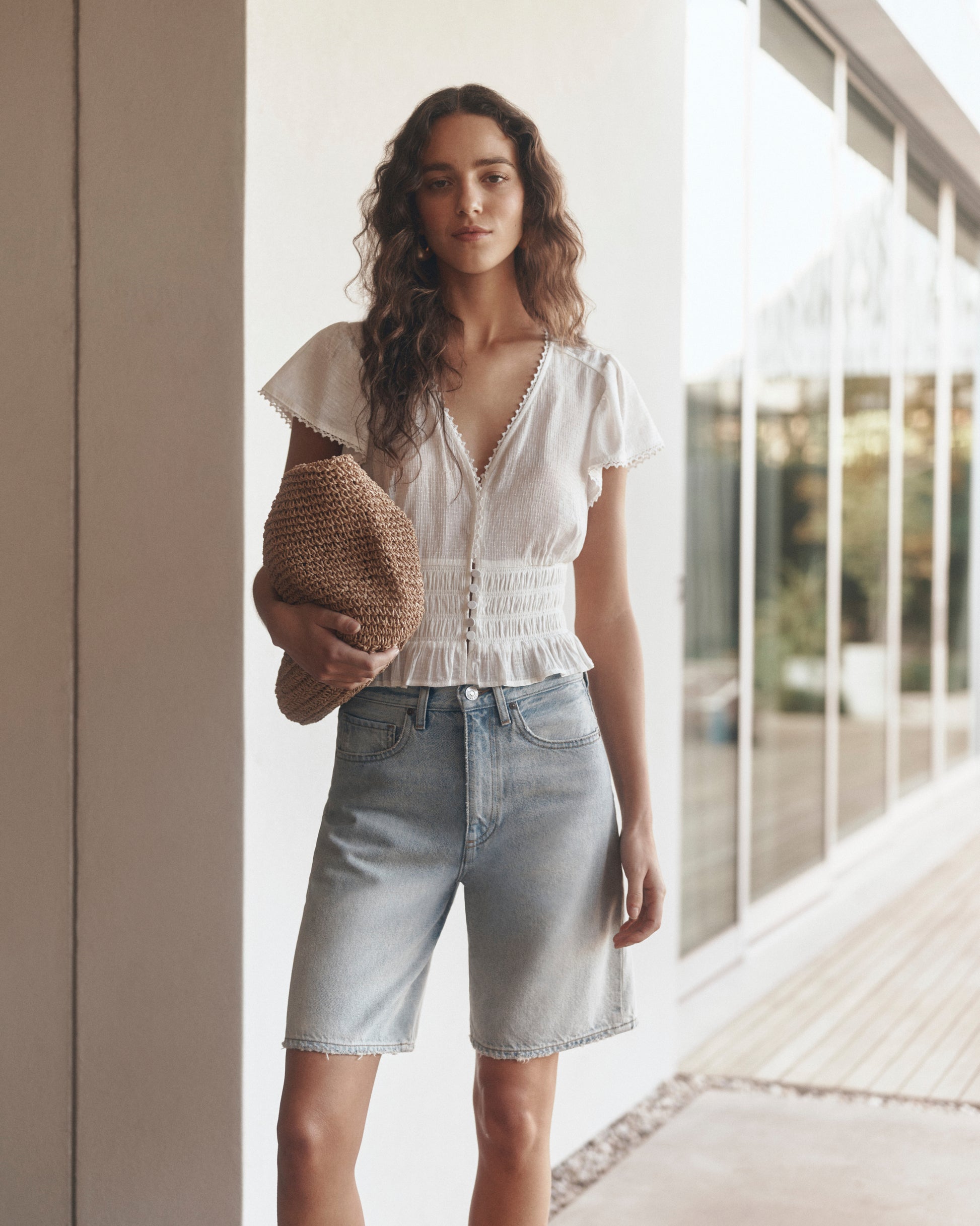 Woman wearing a white blouse and denim shorts holding a brown bag indoors.