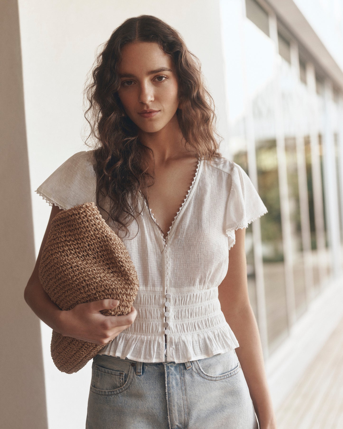 Woman wearing a white blouse and denim shorts holding a woven bag outdoors