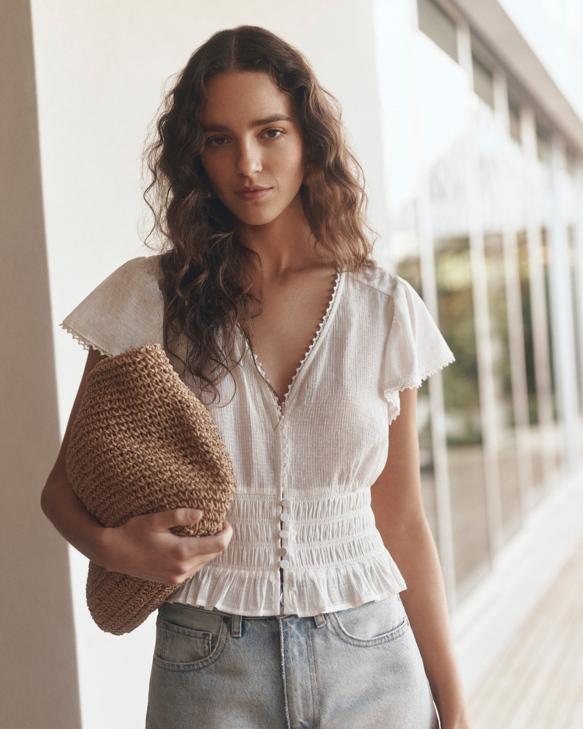 Woman wearing a white blouse and denim shorts holding a woven bag outdoors