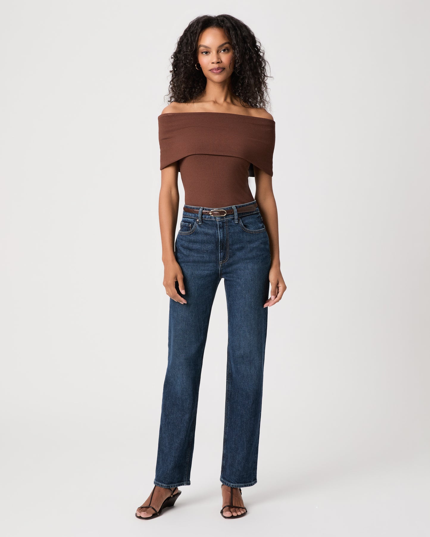 women wearing a brown off the shoulder top and blue jeans paired with brown sandals against a white background 