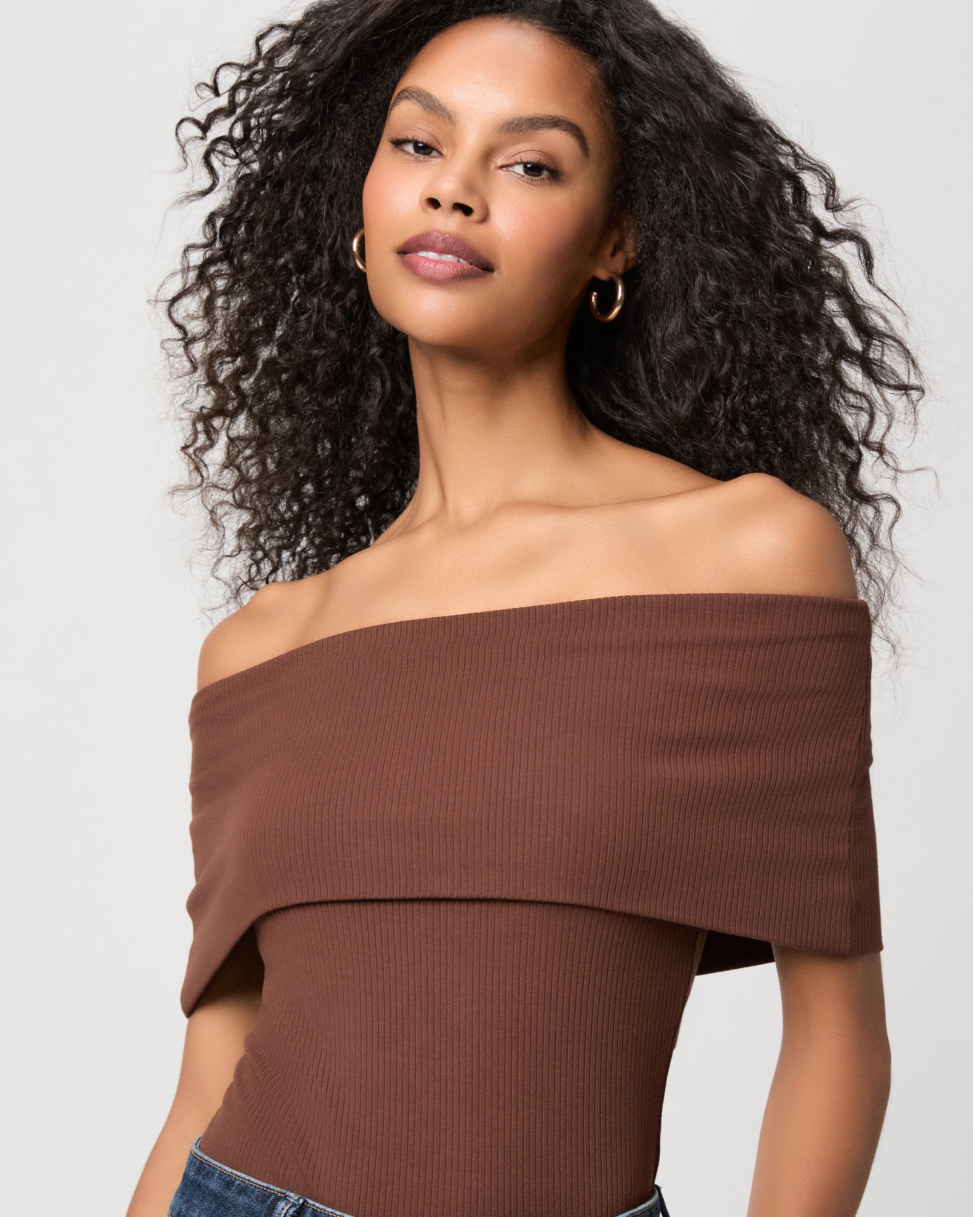 women wearing a brown off the shoulder top and blue jeans against a white background 
