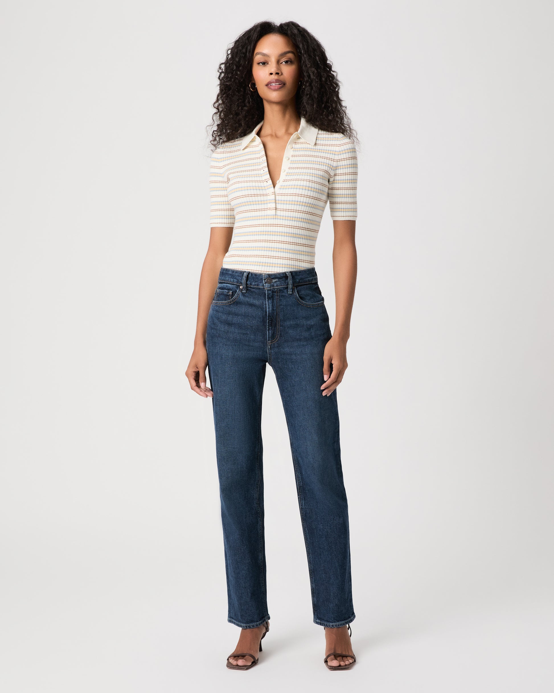 A woman stands against a light gray studio background wearing a beige ribbed tank top, light-wash wide-leg jeans, and brown sandals.
