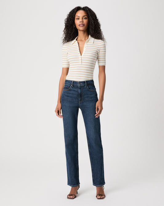 A woman stands against a light gray studio background wearing a beige ribbed tank top, light-wash wide-leg jeans, and brown sandals.