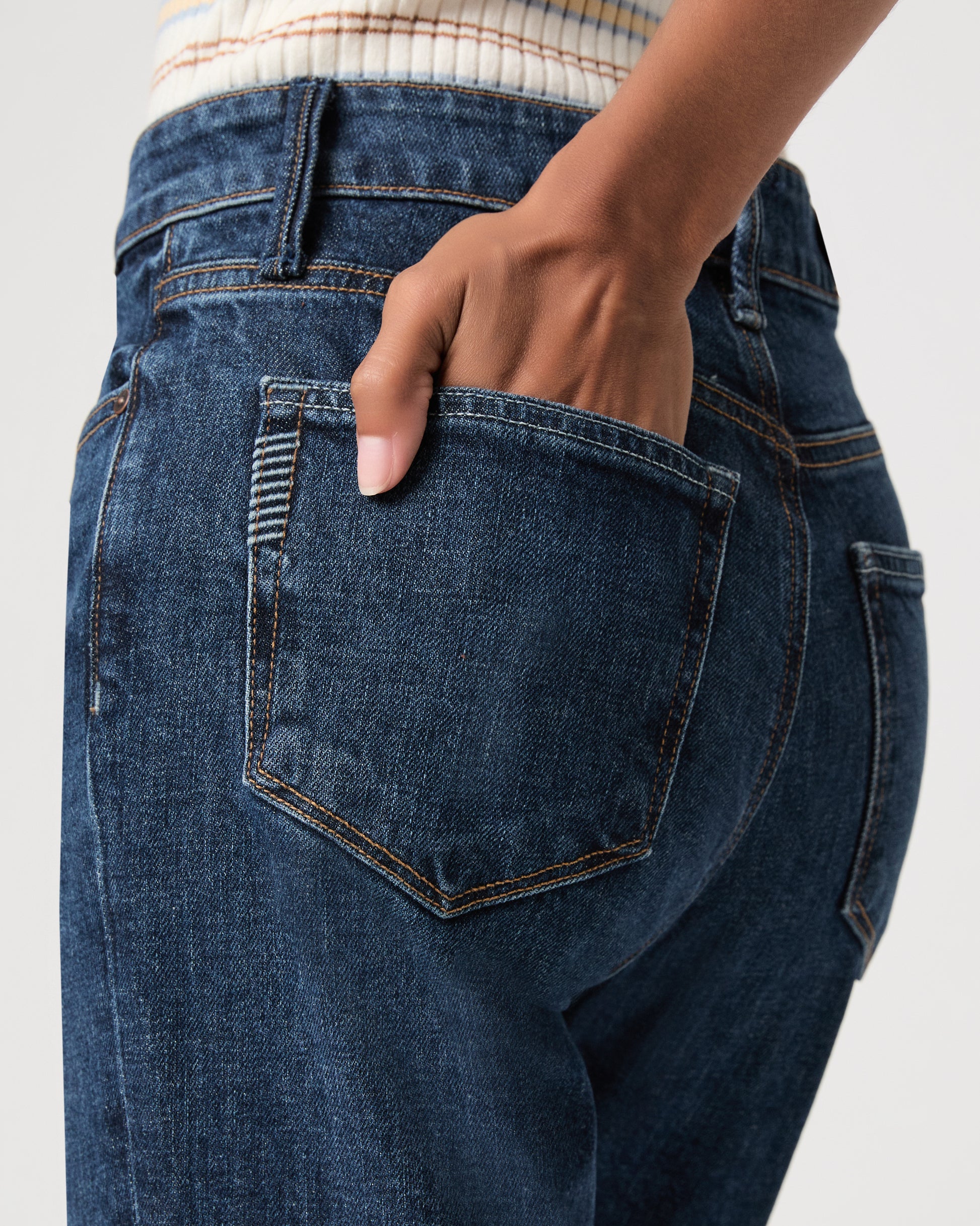 A woman stands against a light gray studio background wearing a beige ribbed tank top, light-wash wide-leg jeans