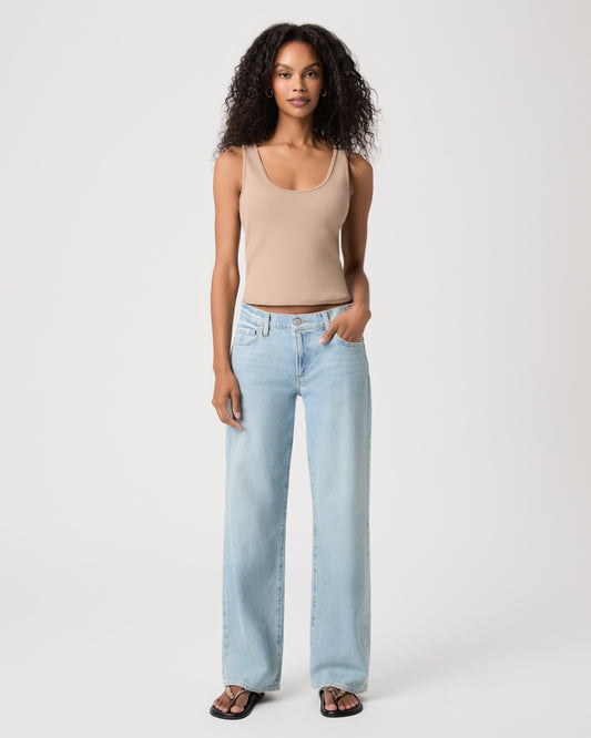 A model stands against a light gray studio background wearing a beige ribbed tank top, light-wash wide-leg jeans, and black sandals.