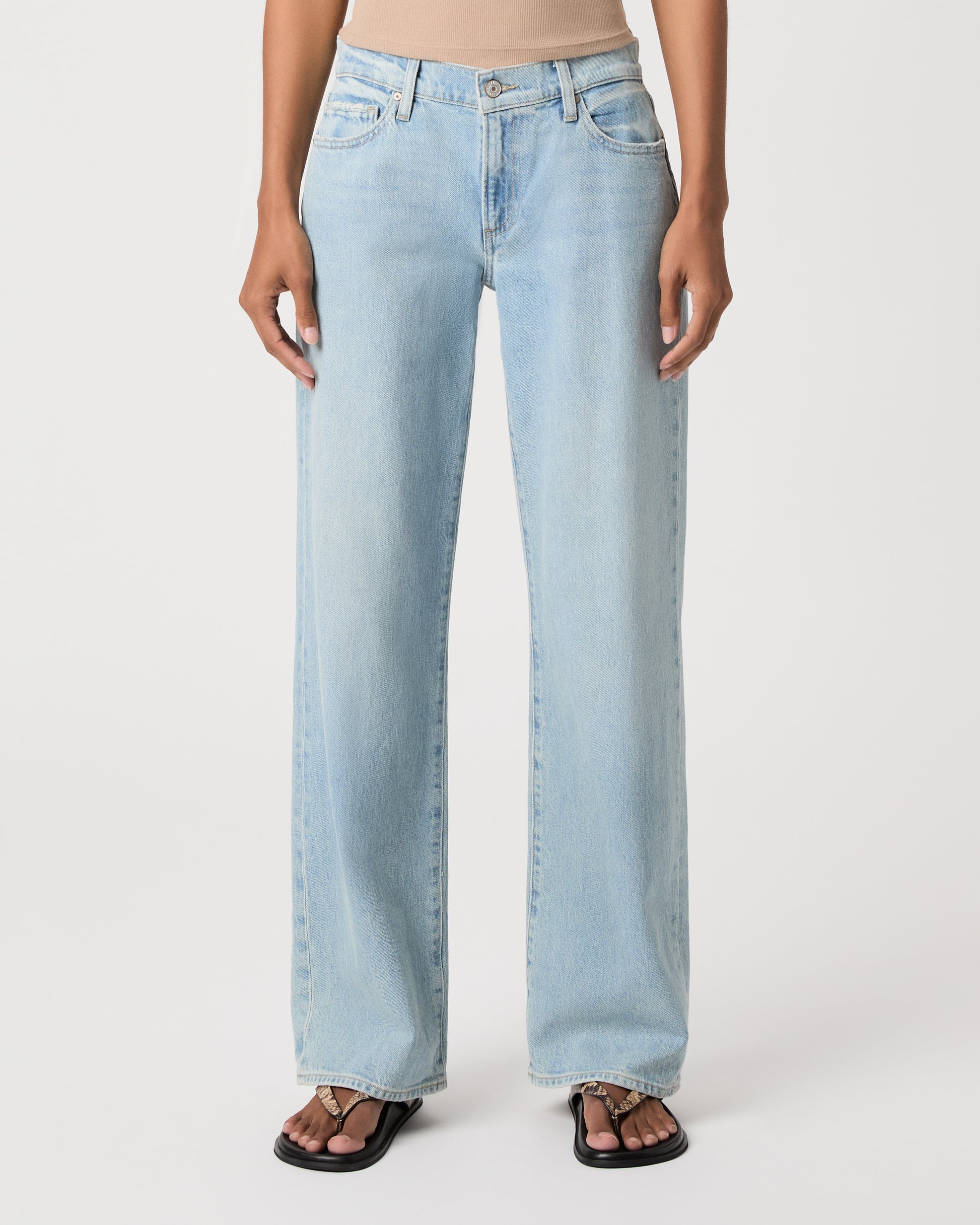 A model stands against a light gray studio background wearing a beige ribbed tank top, light-wash wide-leg jeans, and black sandals.