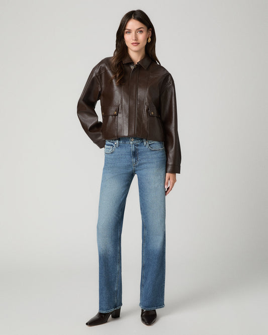A woman modeling a dark brown leather cropped jacket with front flap pockets, paired with blue jeans and black pointed-toe boots, against a light gray studio background.
