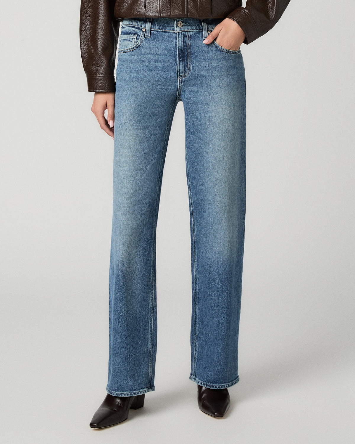A model wearing a pair of medium-wash blue straight-leg jeans, styled with a brown leather jacket and dark brown pointed-toe boots, against a light gray studio background.