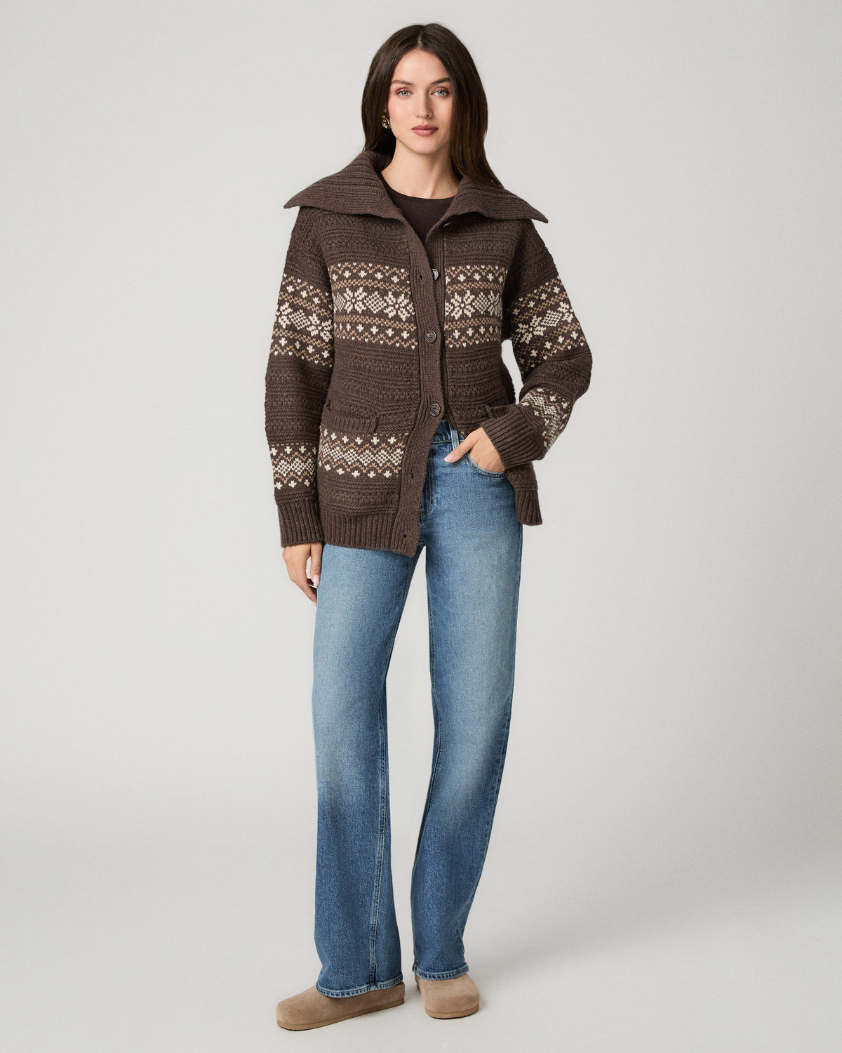 A woman modeling a brown knit cardigan with cream Fair Isle patterns, paired with blue jeans and tan shoes, against a light gray studio background.