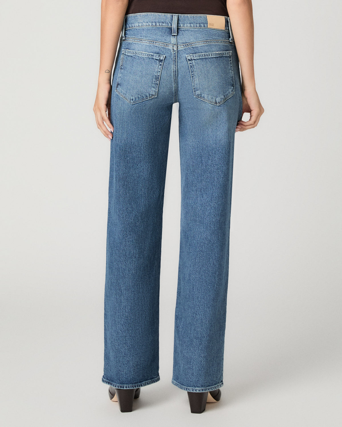 Rear view of a model wearing high-waisted, wide-leg blue denim jeans with a light tan leather patch on the back waistband, paired with black heels against a light gray studio background.