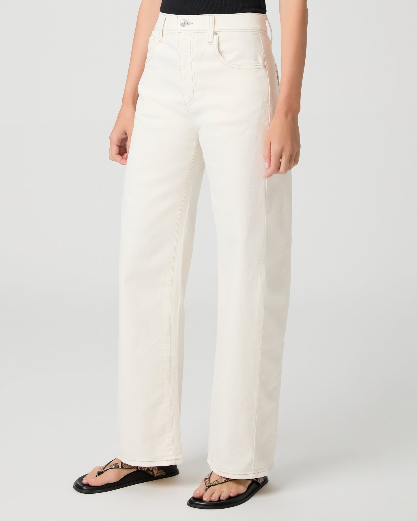 Woman wearing a black tank top and white pants and snake print sandals on a plain background