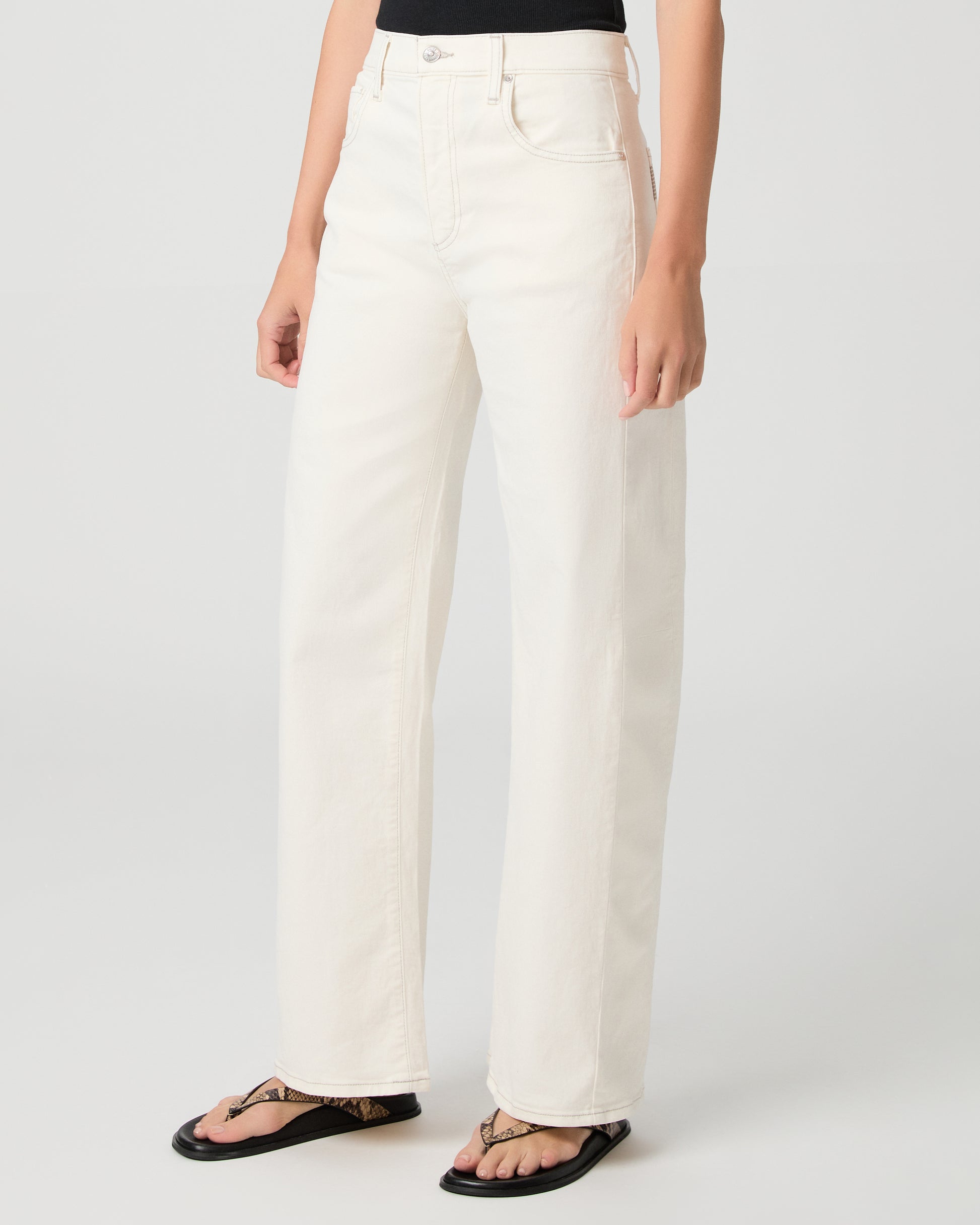 Woman wearing a black tank top and white pants and snake print sandals on a plain background