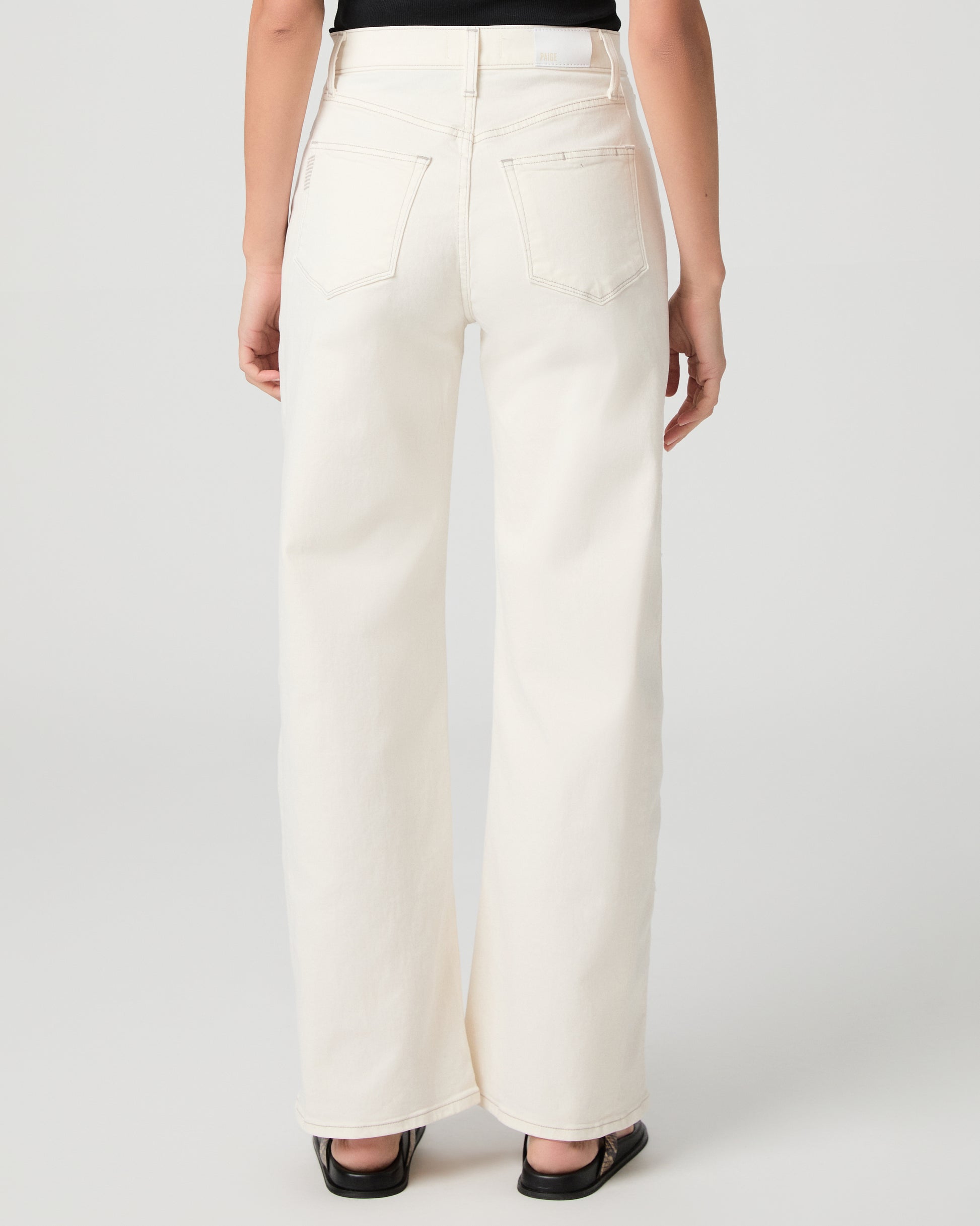 Woman wearing a black tank top and white pants and snake print sandals on a plain background