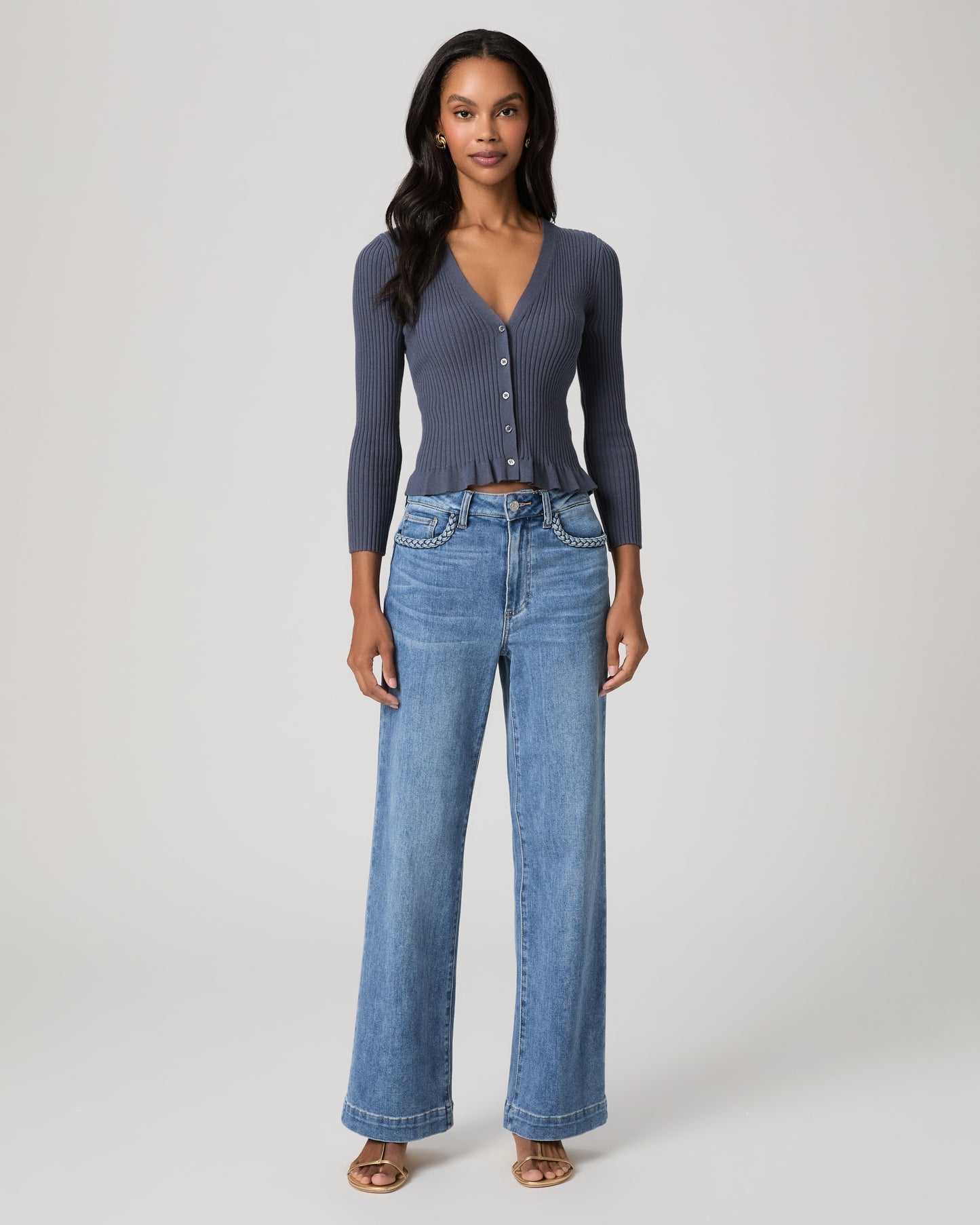 Woman wearing a blue long sleeve top and blue jeans and gold sandals on a white background