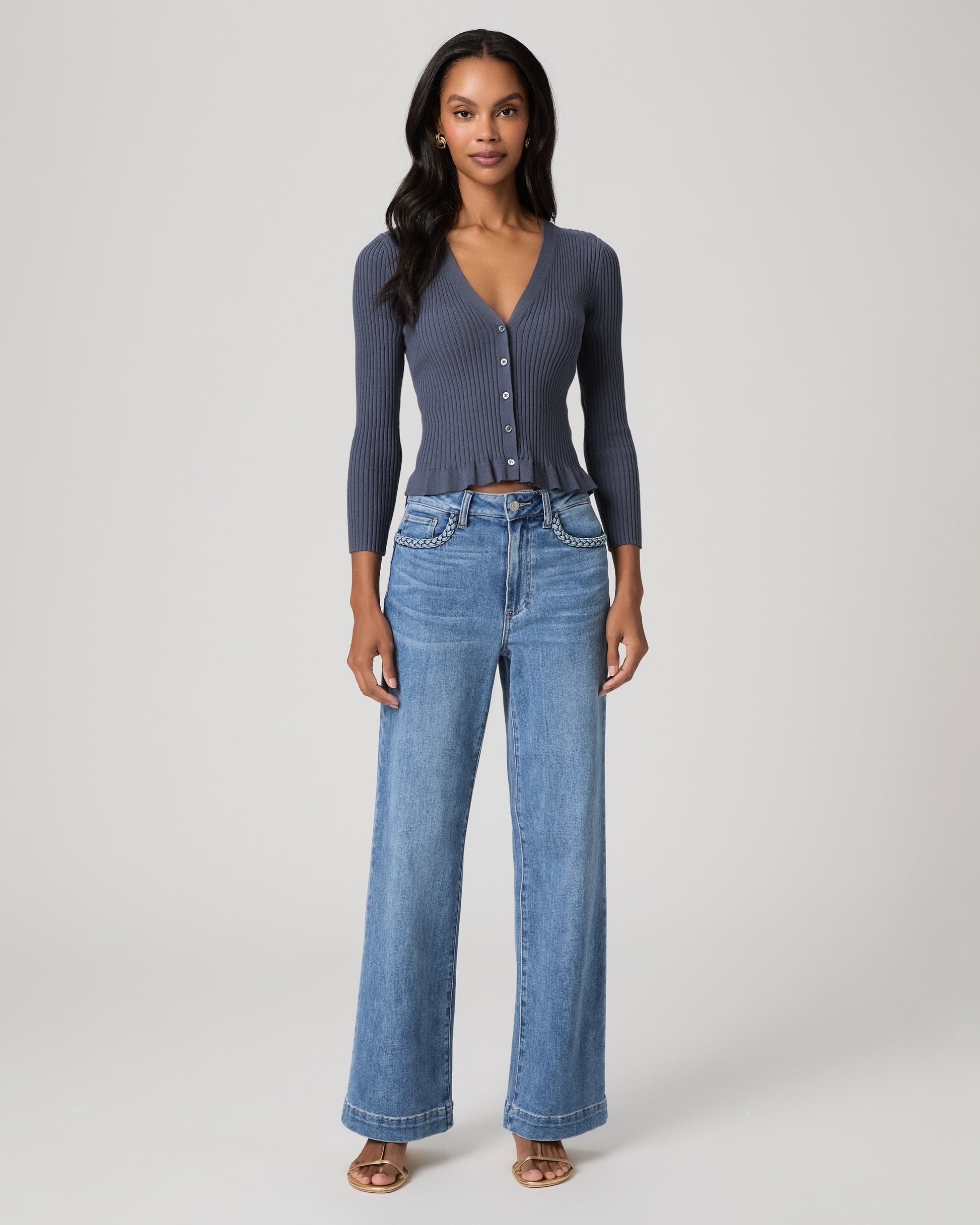 Woman wearing a blue long sleeve top and blue jeans and gold sandals on a white background