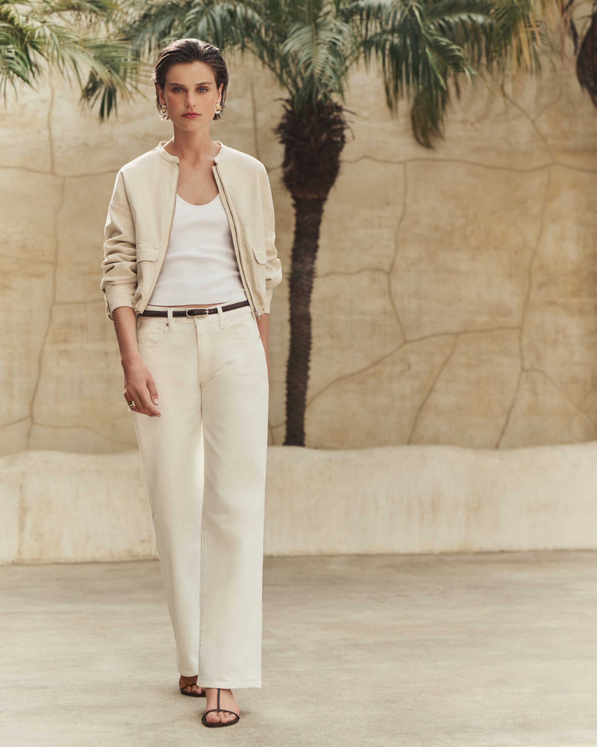 Woman in a beige jacket and white pants standing in front of a textured wall with palm trees.