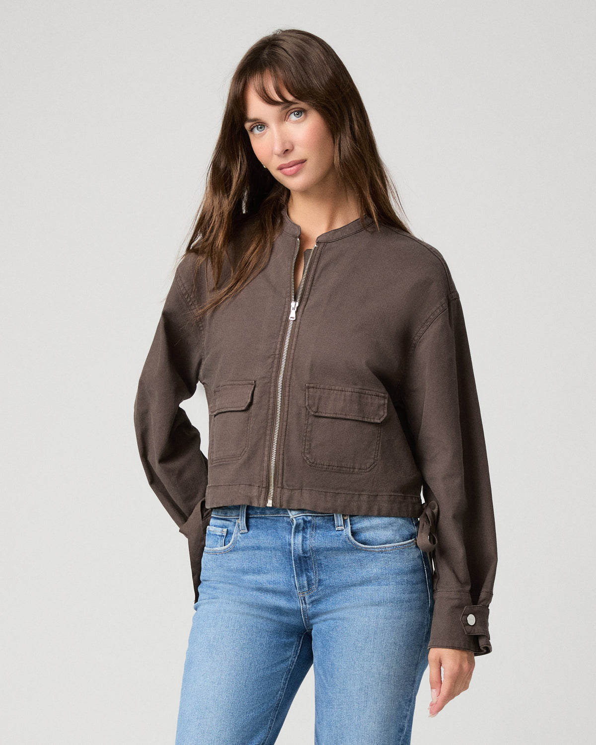 Woman wearing brown jacket and blue pants with a white background.
