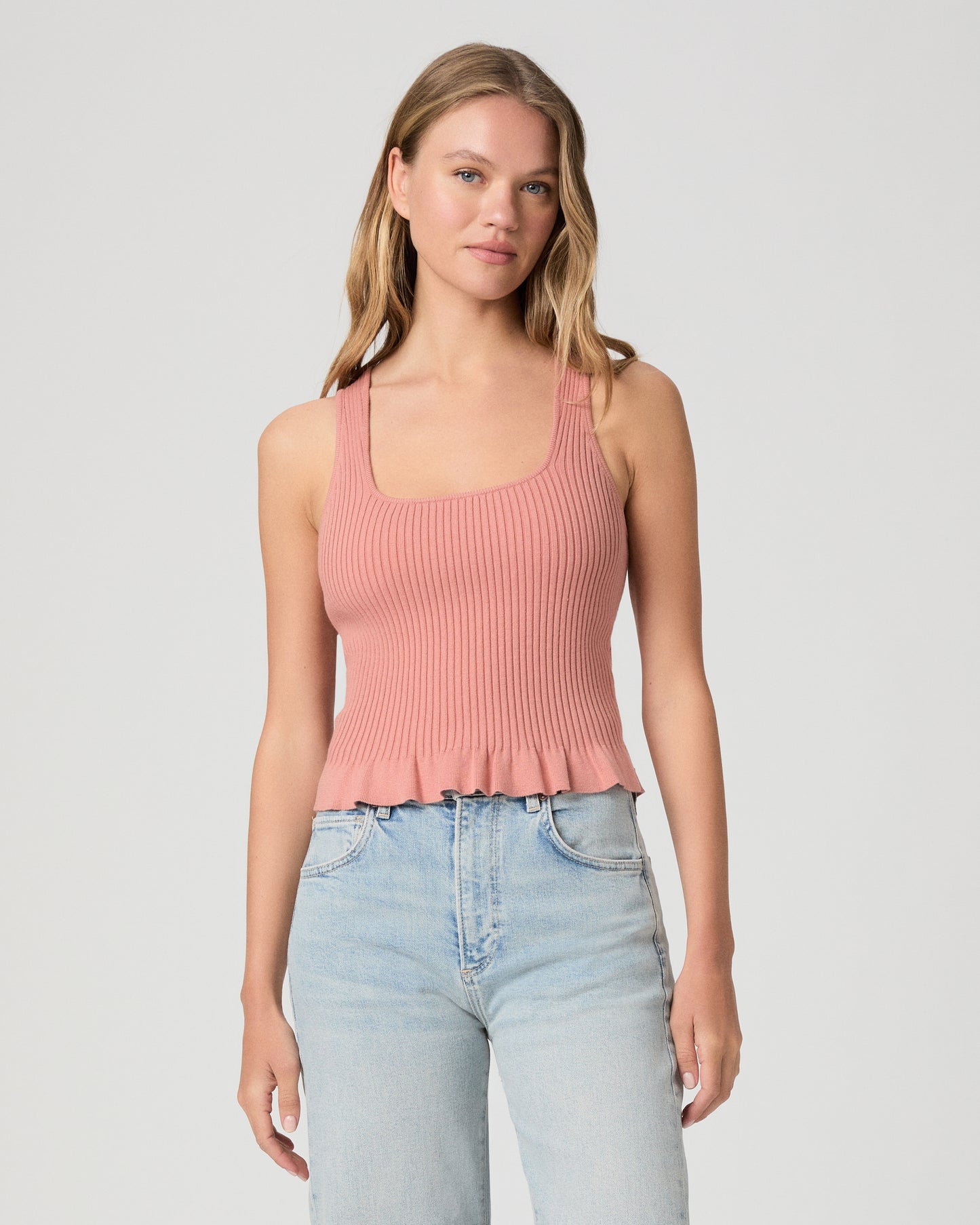 Woman wearing a pink ribbed tank top and light blue jeans on a plain background