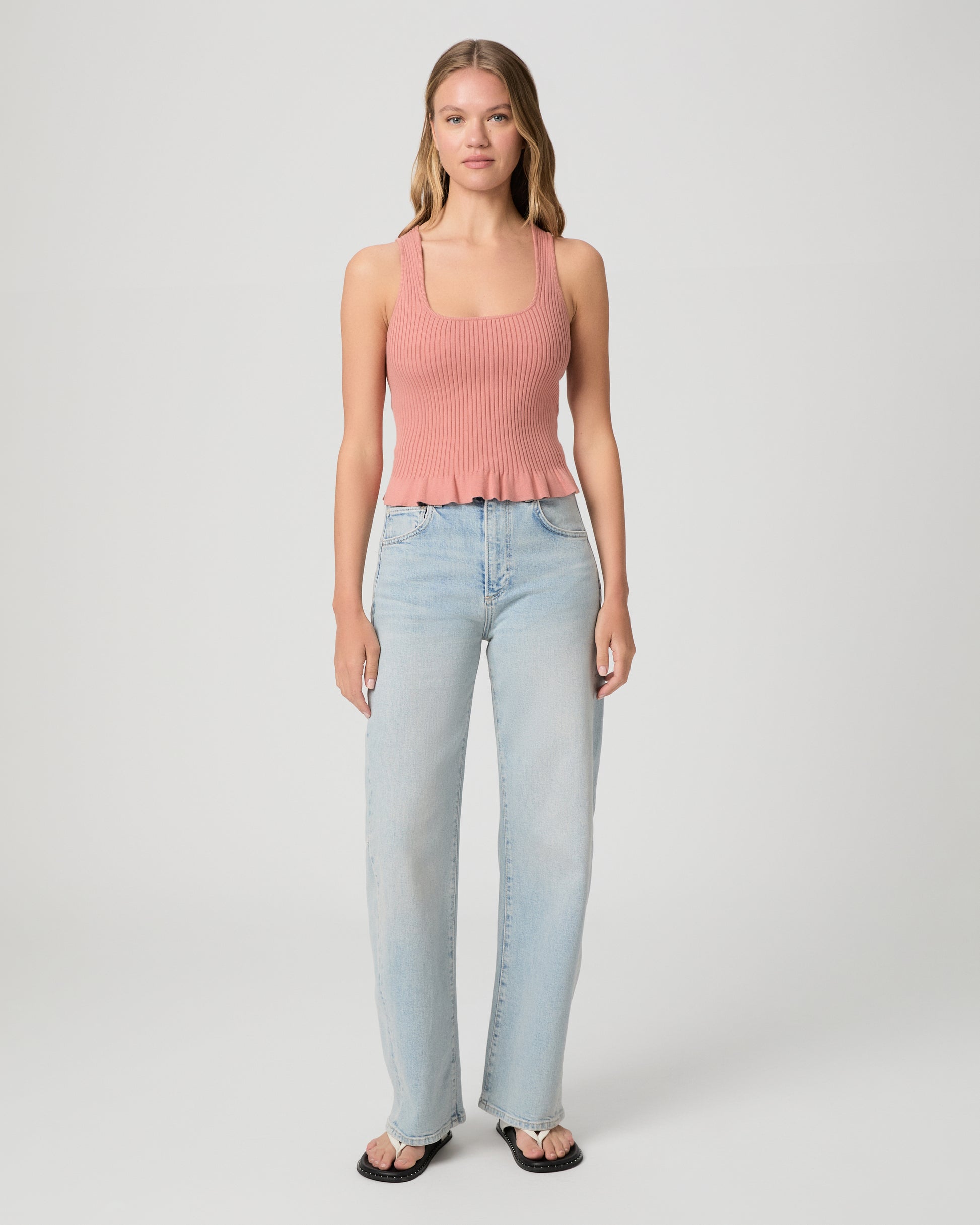 Woman wearing a pink ribbed tank top and light blue jeans on a plain background