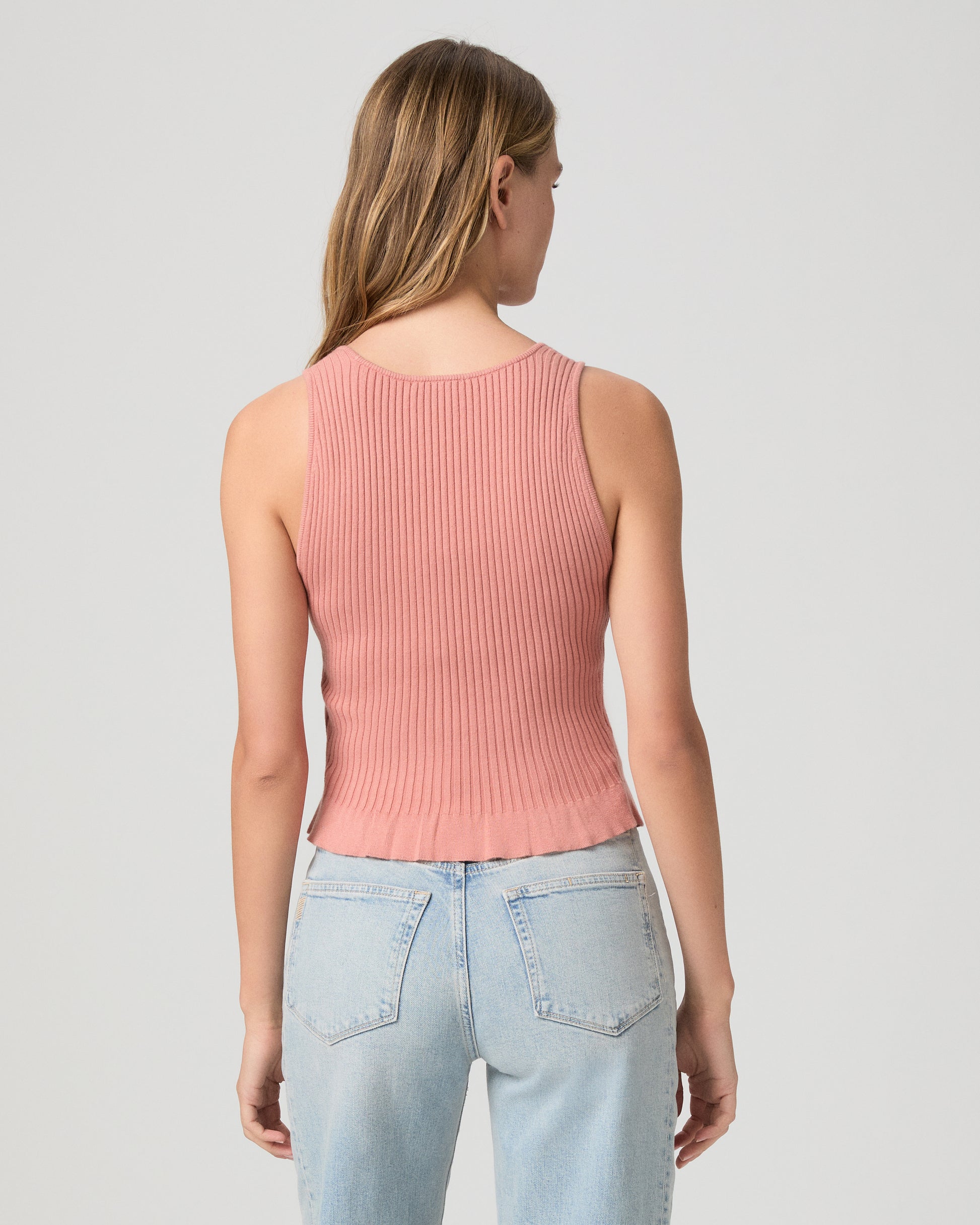 Woman wearing a pink ribbed tank top and light blue jeans on a plain background
