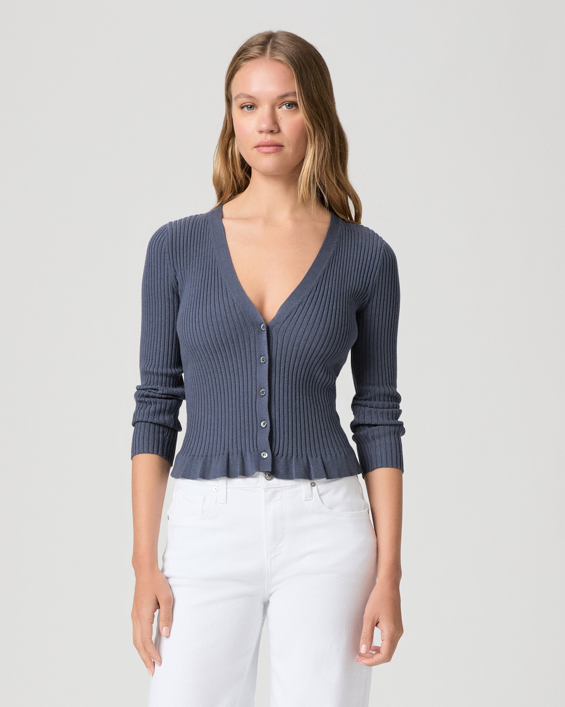 Woman wearing a blue ribbed cardigan and white pants on a light gray background