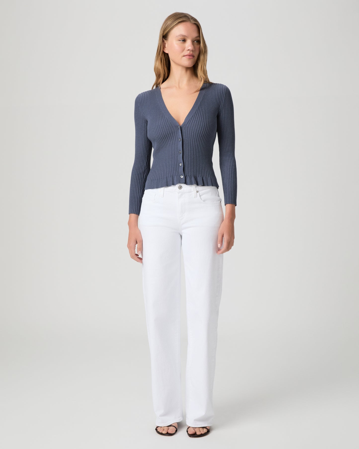 Woman wearing a blue ribbed cardigan and white pants on a light gray background