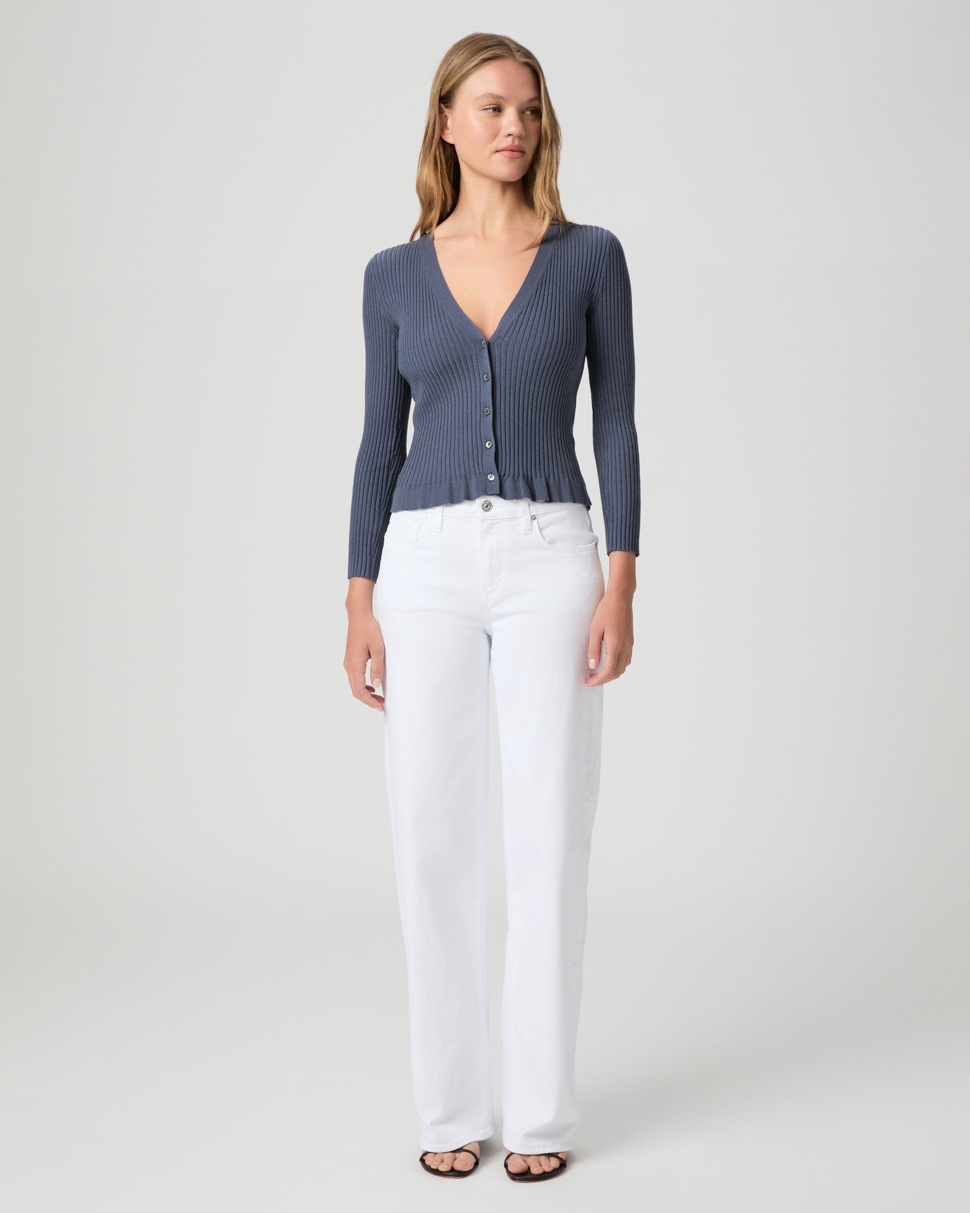 Woman wearing a blue ribbed cardigan and white pants on a light gray background