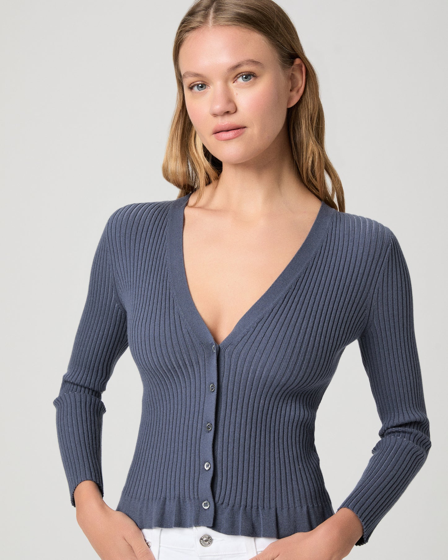 Woman wearing a blue ribbed cardigan and white pants on a light gray background